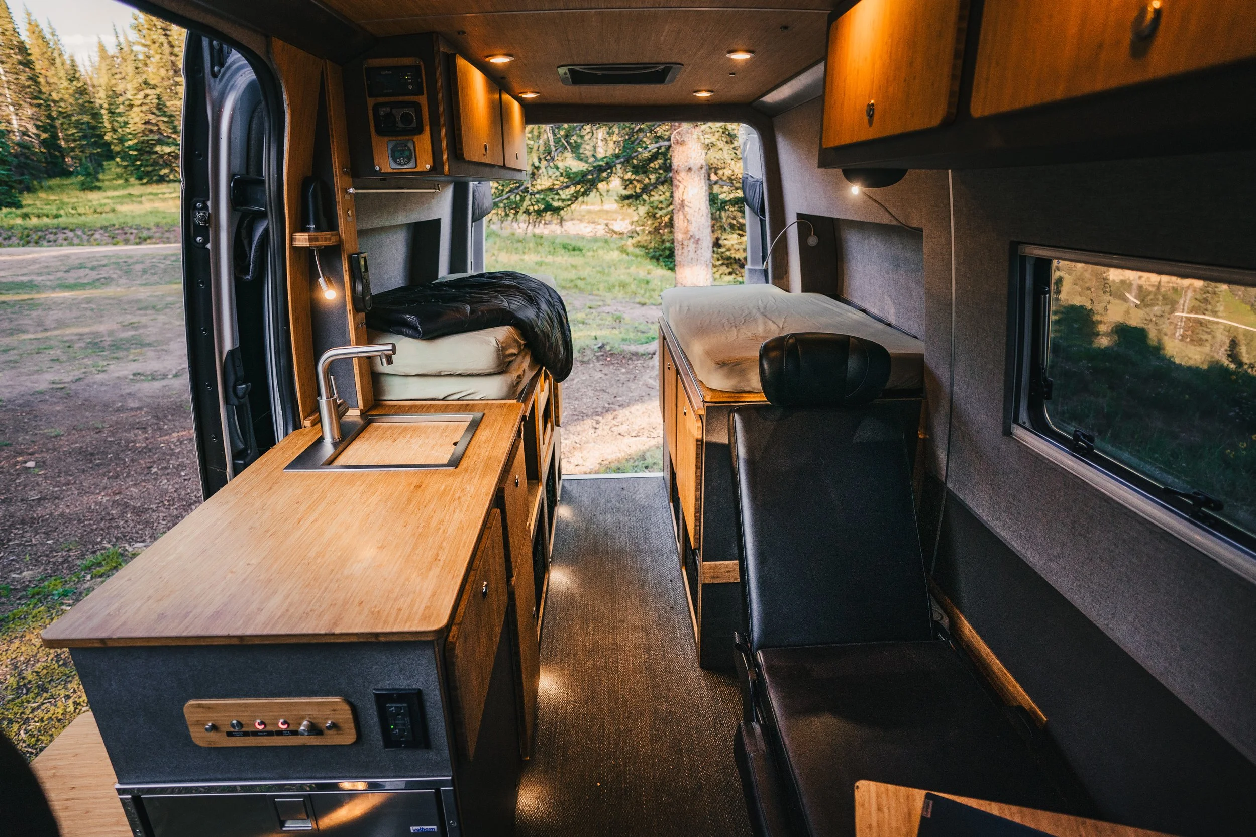 Interior of a camper van with wooden cabinets, a sink, a bench seat, and a bed, with a view of trees outside the window.