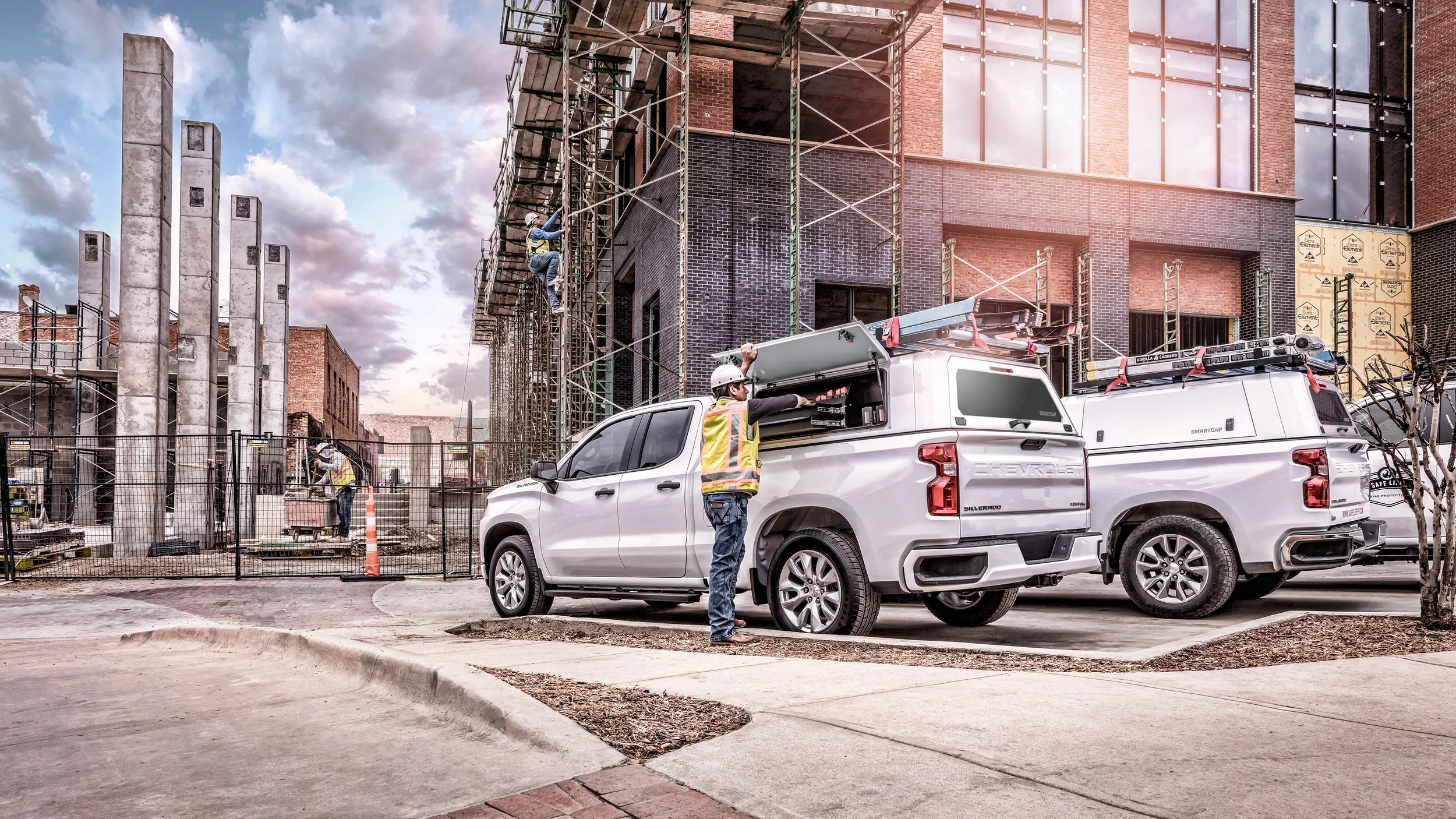 Construction workers working on a building site with scaffolding. Two white pickup trucks parked in the foreground, with tools stored in the truck beds. The building under construction has brick and glass walls. The sky is partly cloudy.