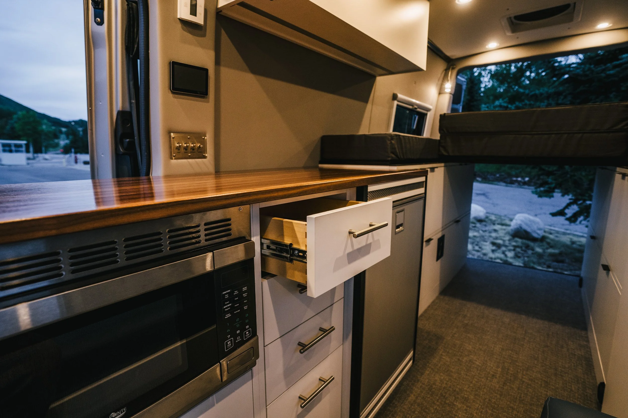 Interior of a camper van, showing a kitchenette with a microwave, cabinets, and a countertop, with a sleeping area above and a view outside through the open door.