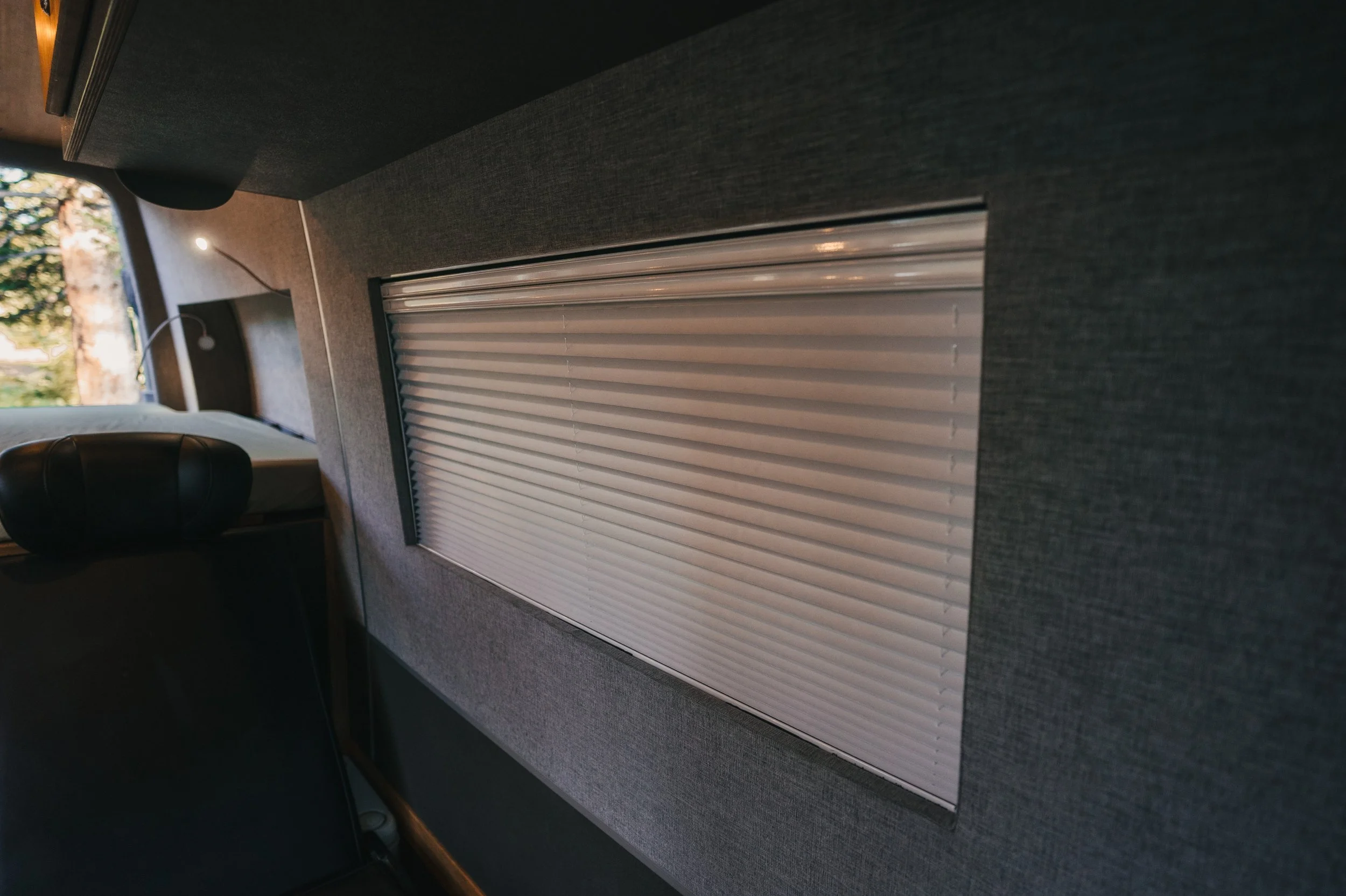 Interior of a vehicle with a closed window with a white blind, a black headrest, a small white reading lamp, and a mountain scene visible through the window.