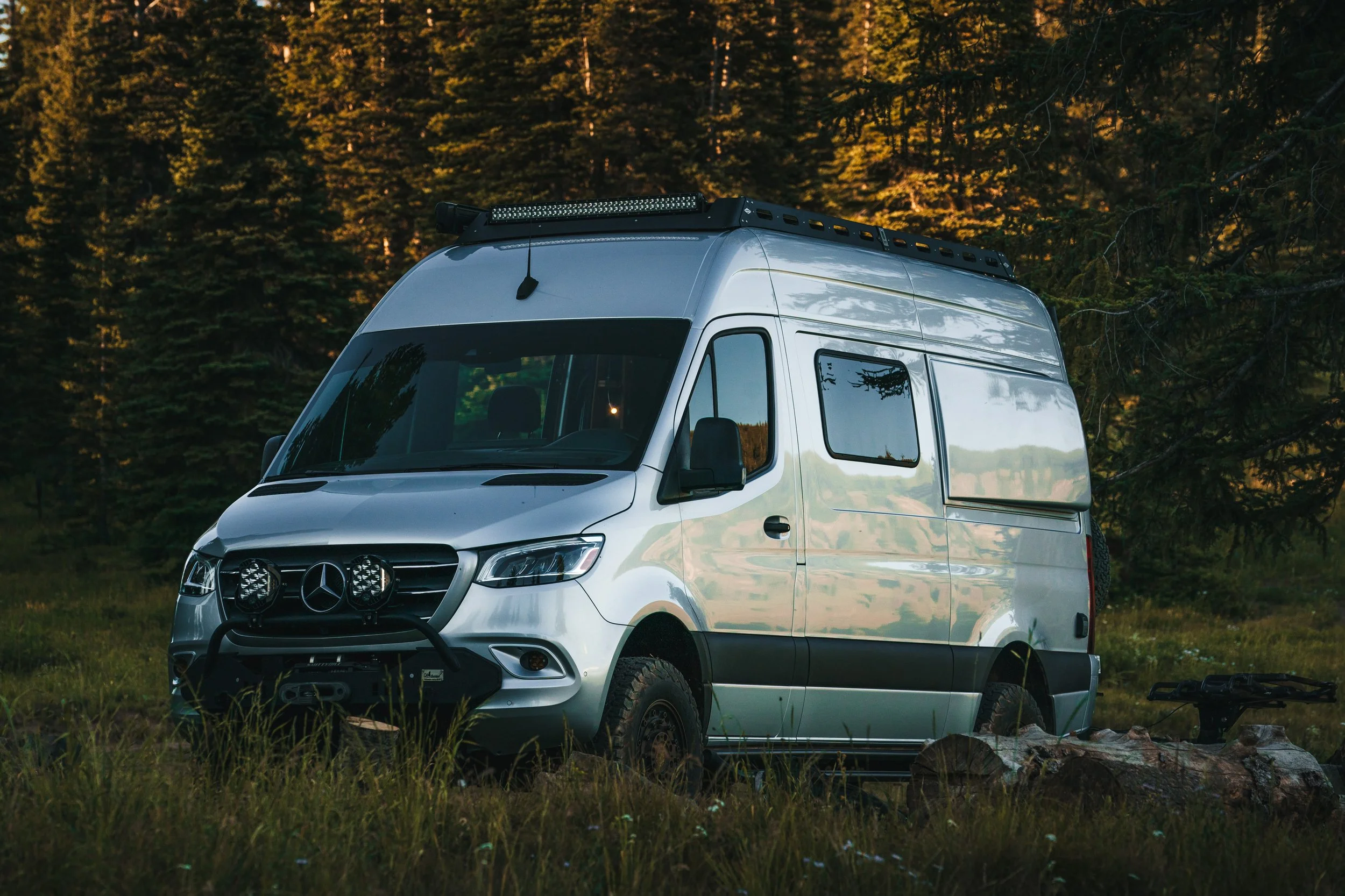 Silver camper van parked in a grassy area with trees in the background at sunset.