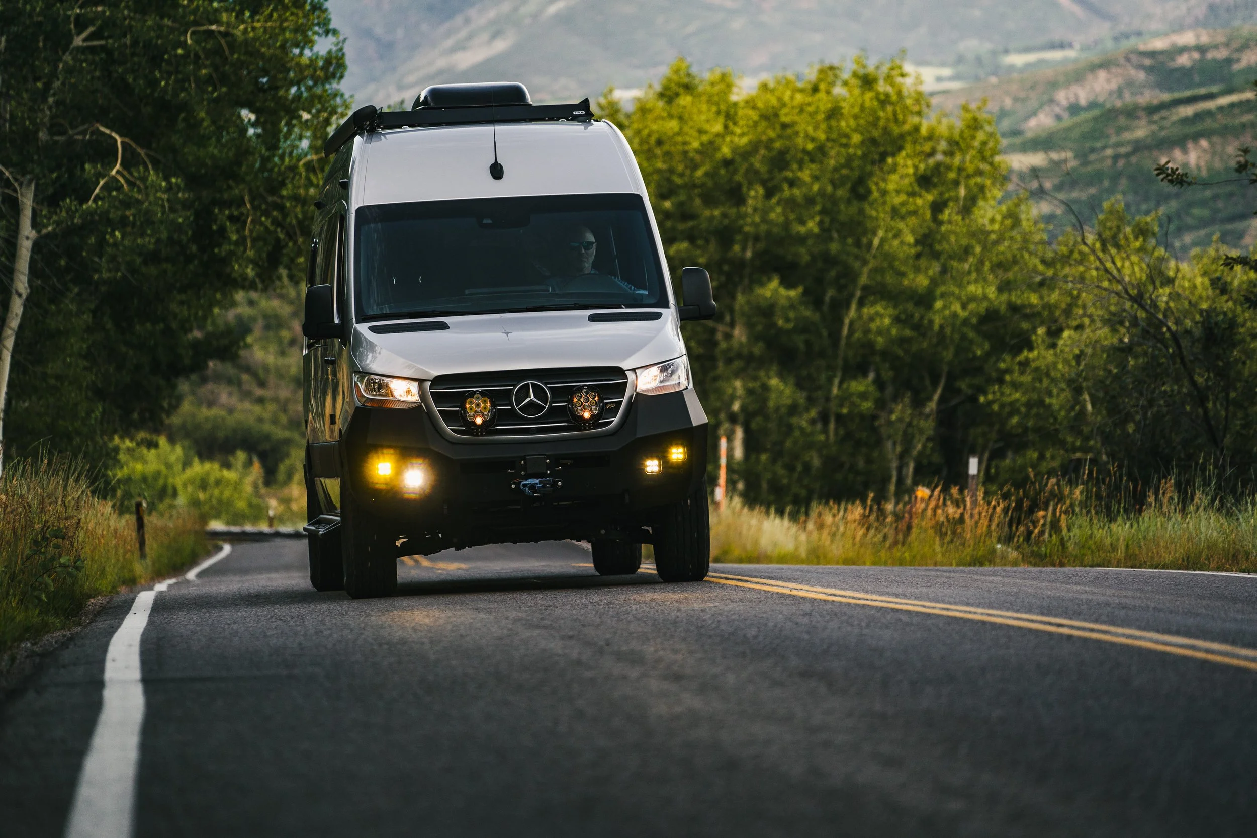 A silver Mercedes-Benz van driving on a winding rural road surrounded by green trees and mountains in the background.