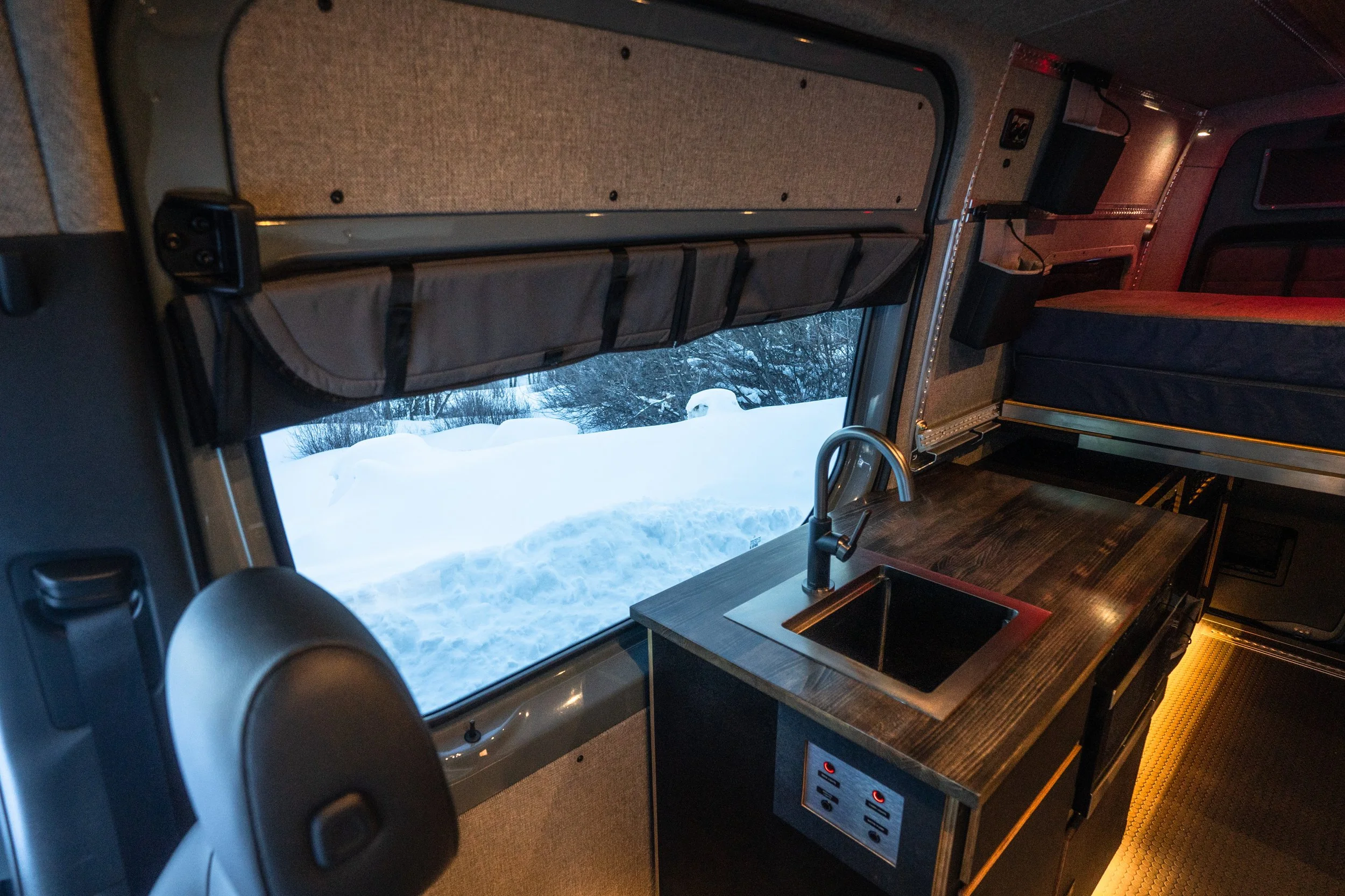 Inside a camper van with a wooden counter, small sink, window showing snow outside, and a bed in the background.