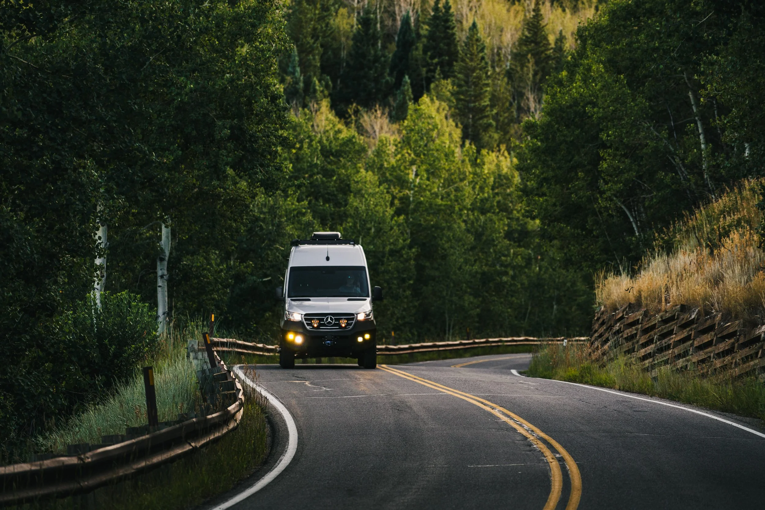 A silver Sprinter van driving on a winding mountain road surrounded by greenery and trees.