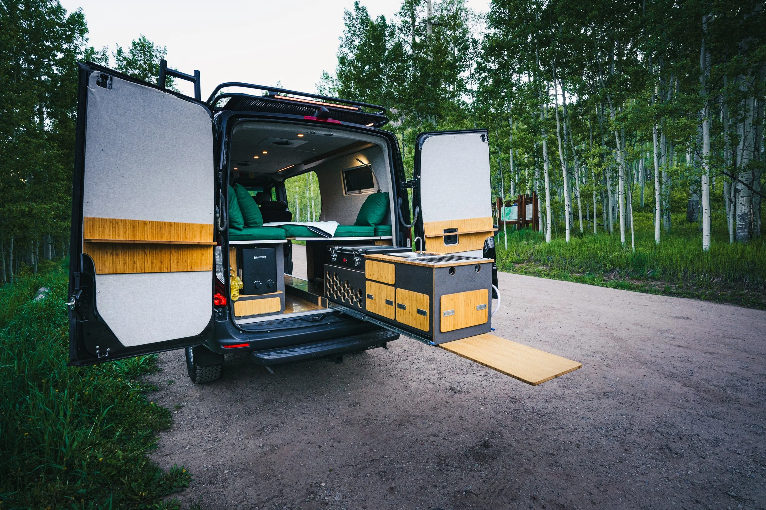 A camper van parked in a forest with its rear doors open, showing a compact living space with green cushions, kitchen area, and a slide-out extension.