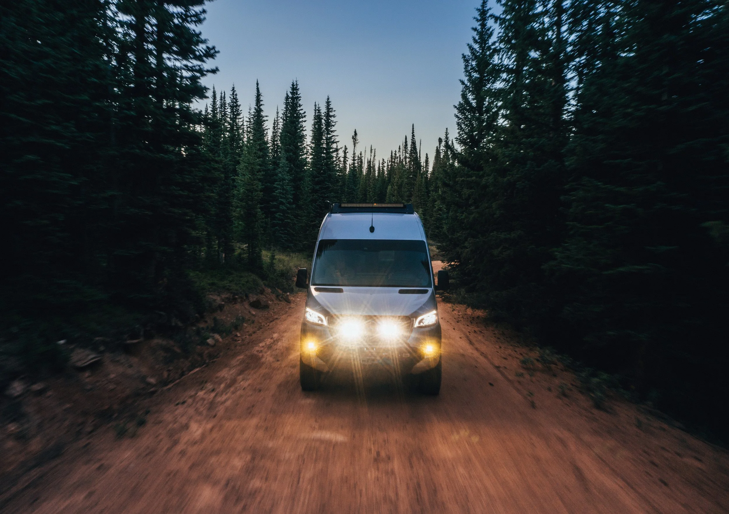 A white van driving on a dirt road through a forested area with tall pine trees, during dusk or early evening.