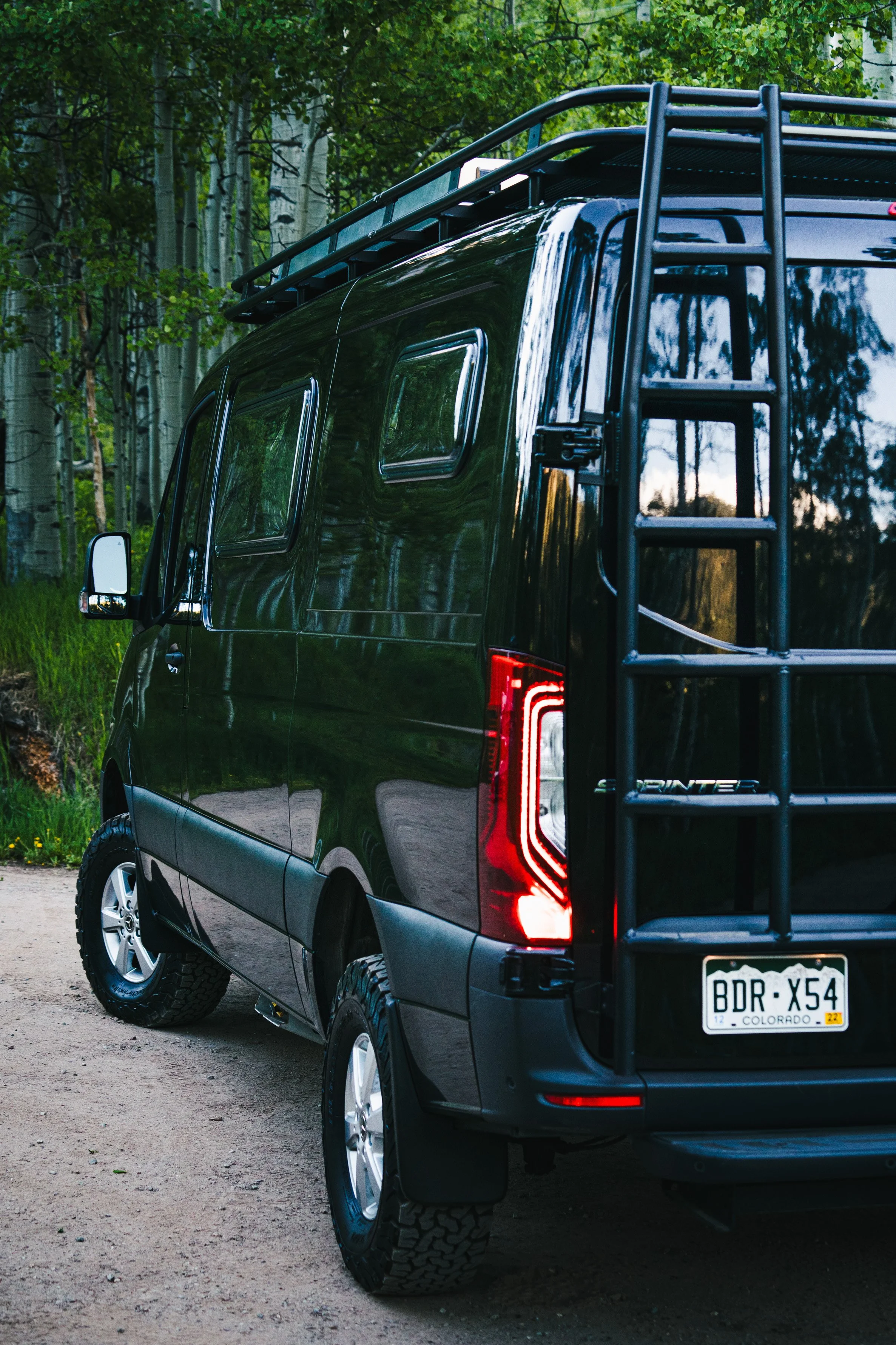 Black camper van parked on a dirt road in a forested area with tall trees and green leaves.