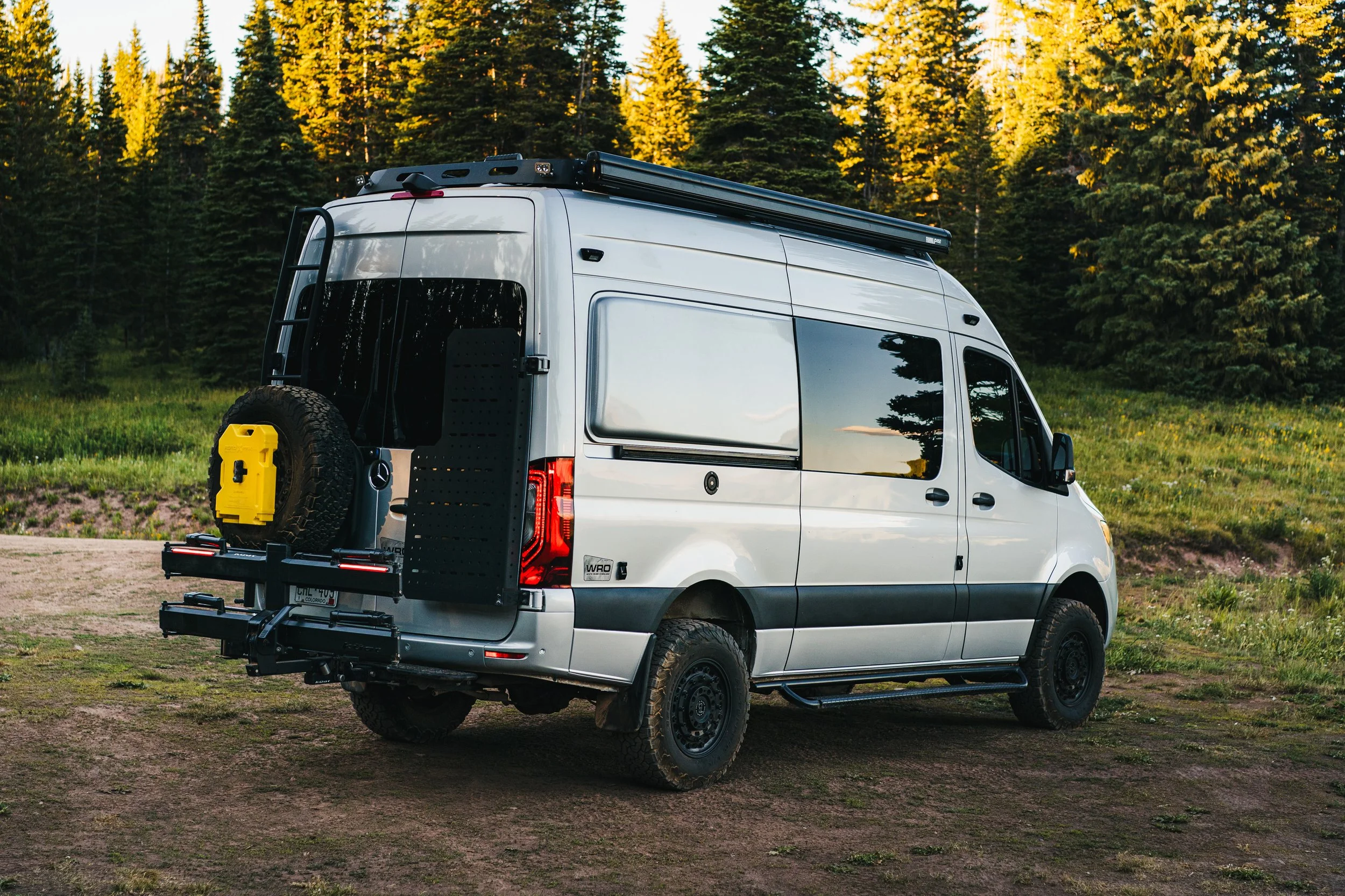 White camper van parked on a dirt path in a forested area with tall pine trees, capturing the rear and side view, with a spare tire mounted on the back.