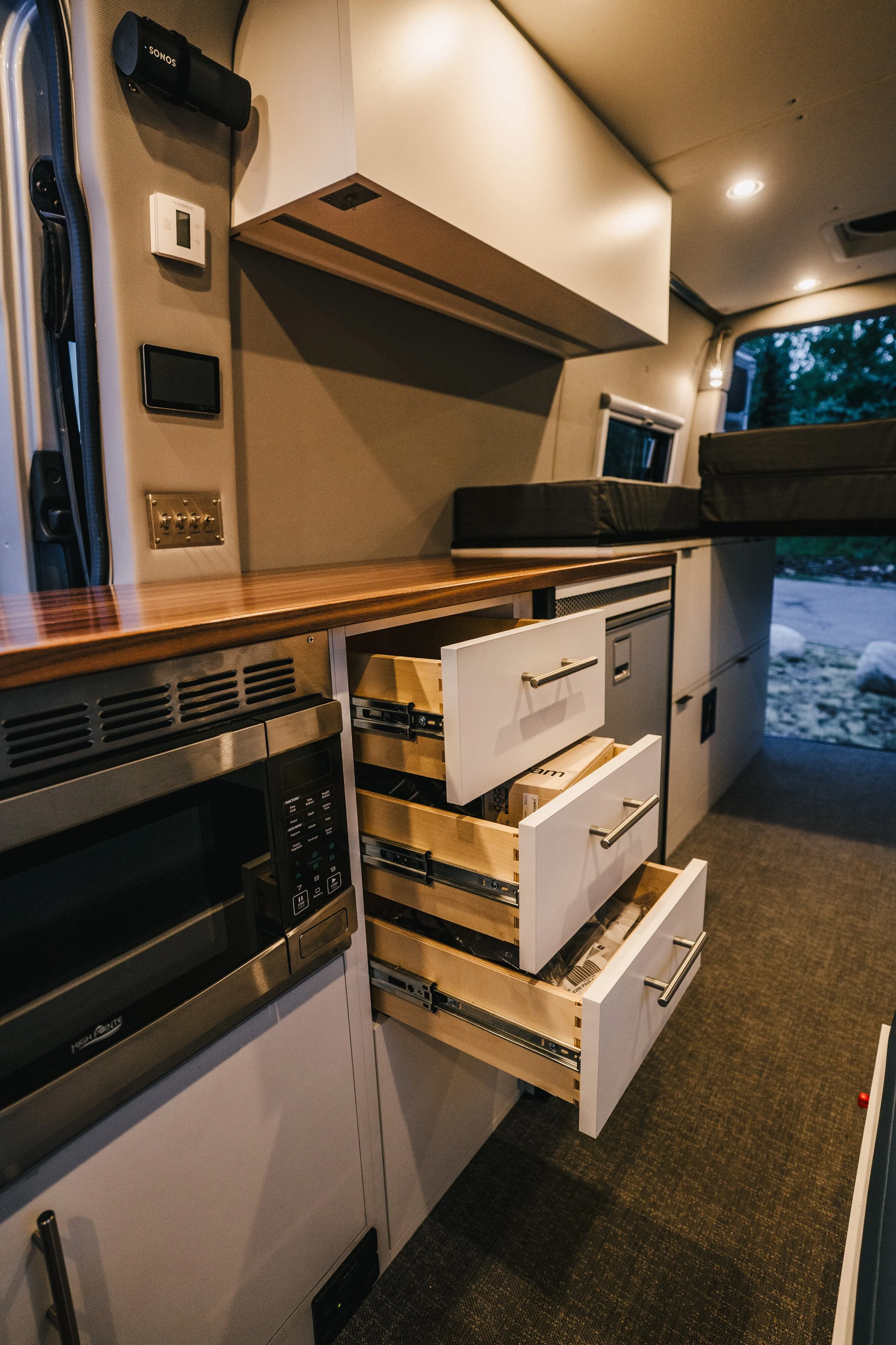Inside a modern camper van with open kitchen drawers, a microwave, and a countertop, with beds and windows visible in the background.