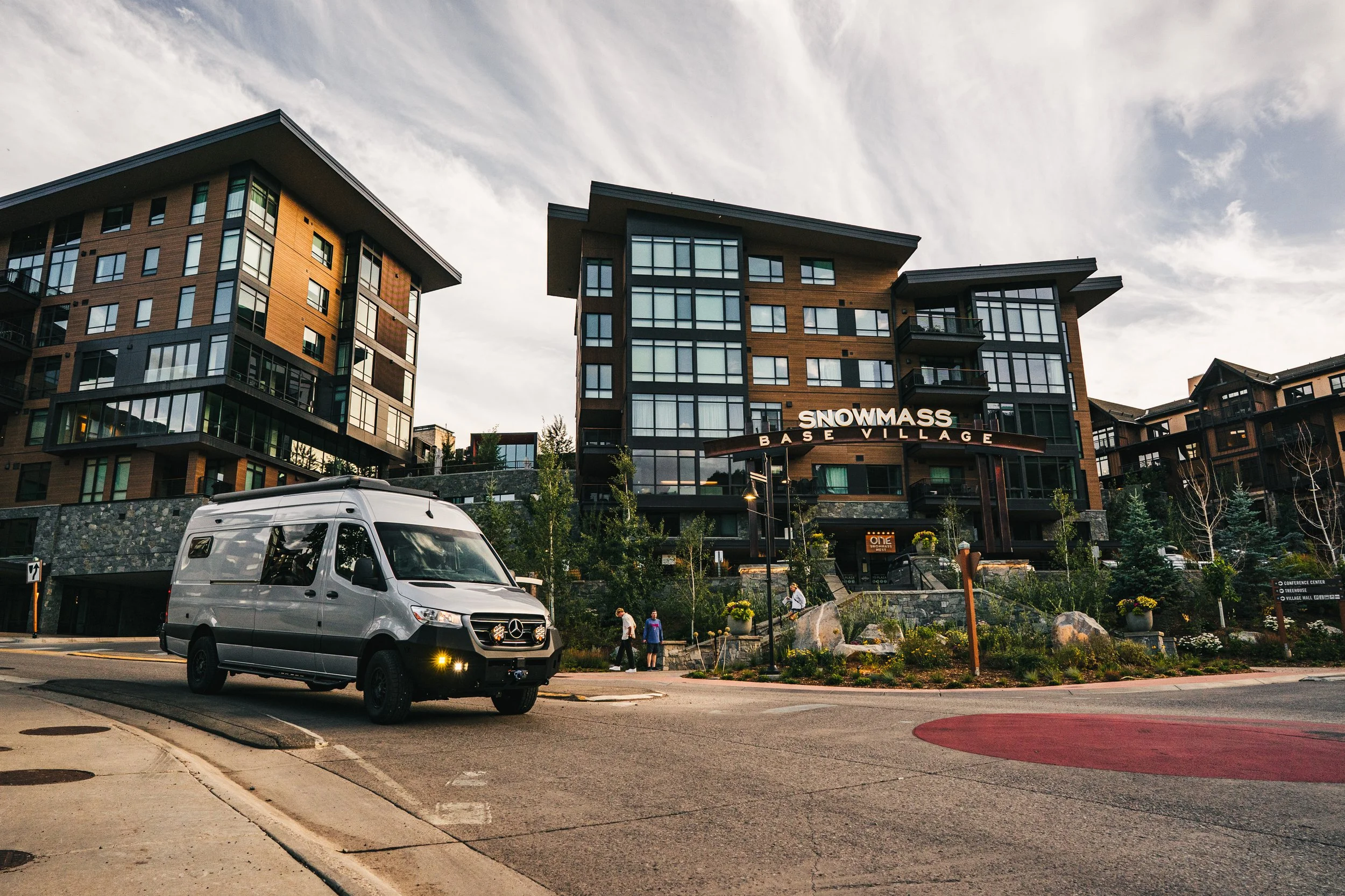 Modern apartment buildings with a parking area and a Mercedes-Benz van in the foreground, under a partly cloudy sky.