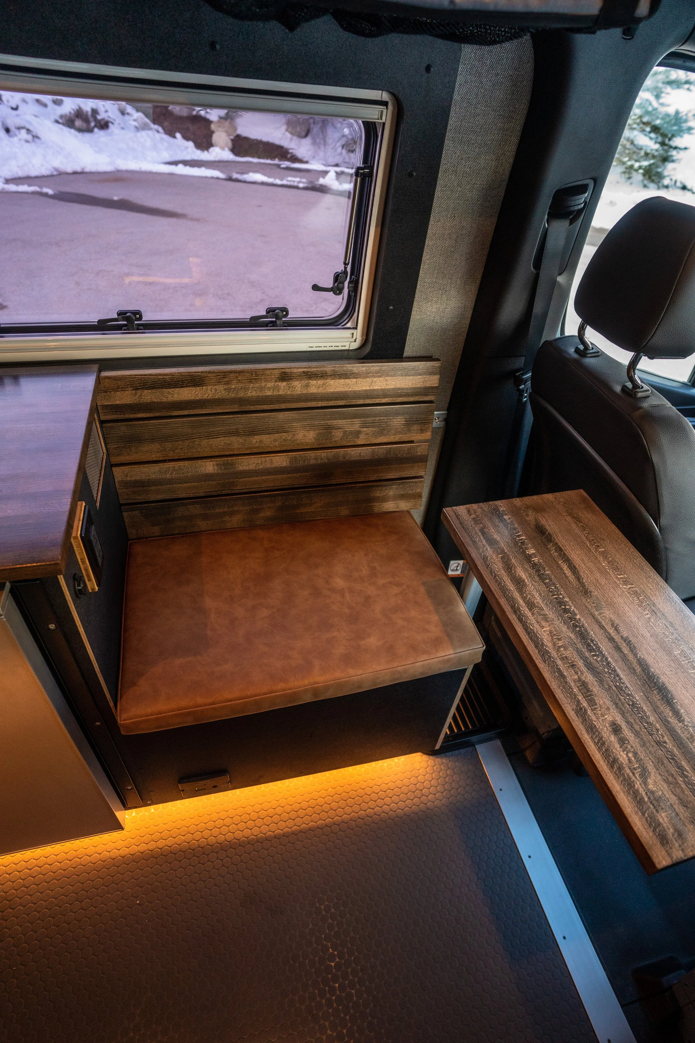 Interior of a camper van showing a small seating area with a brown leather cushion, wooden tables, and a window with snow outside.