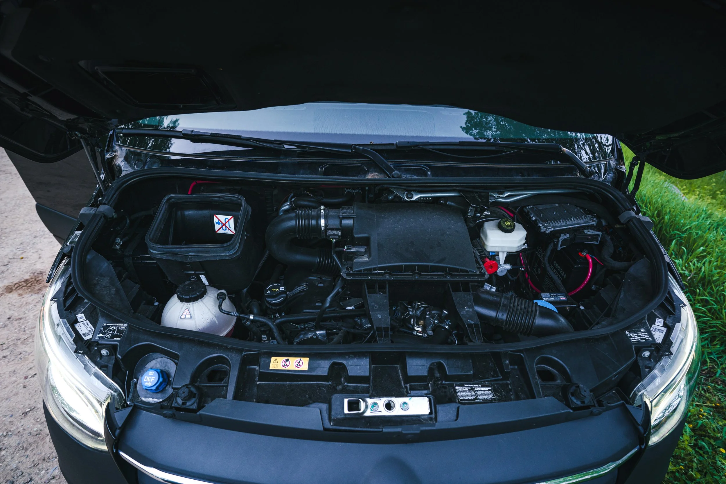Open car hood showing engine components, including air filter, coolant reservoir, and battery, with a grassy background.