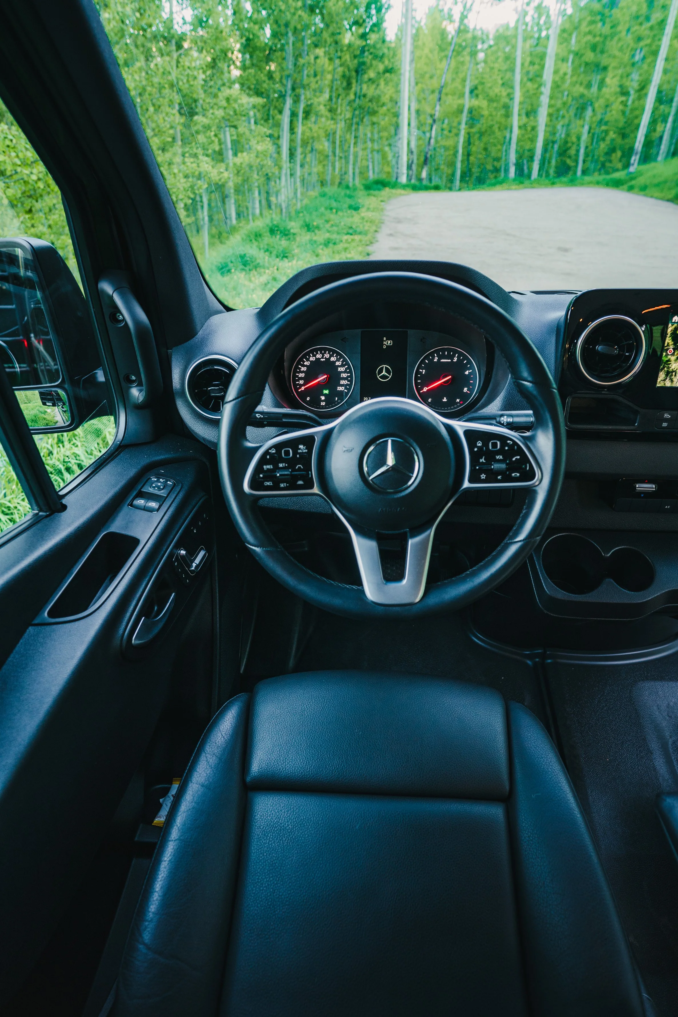The interior of a Mercedes-Benz vehicle, showing the steering wheel, dashboard, and driver's seat, with a view of a green, wooded landscape through the windshield.