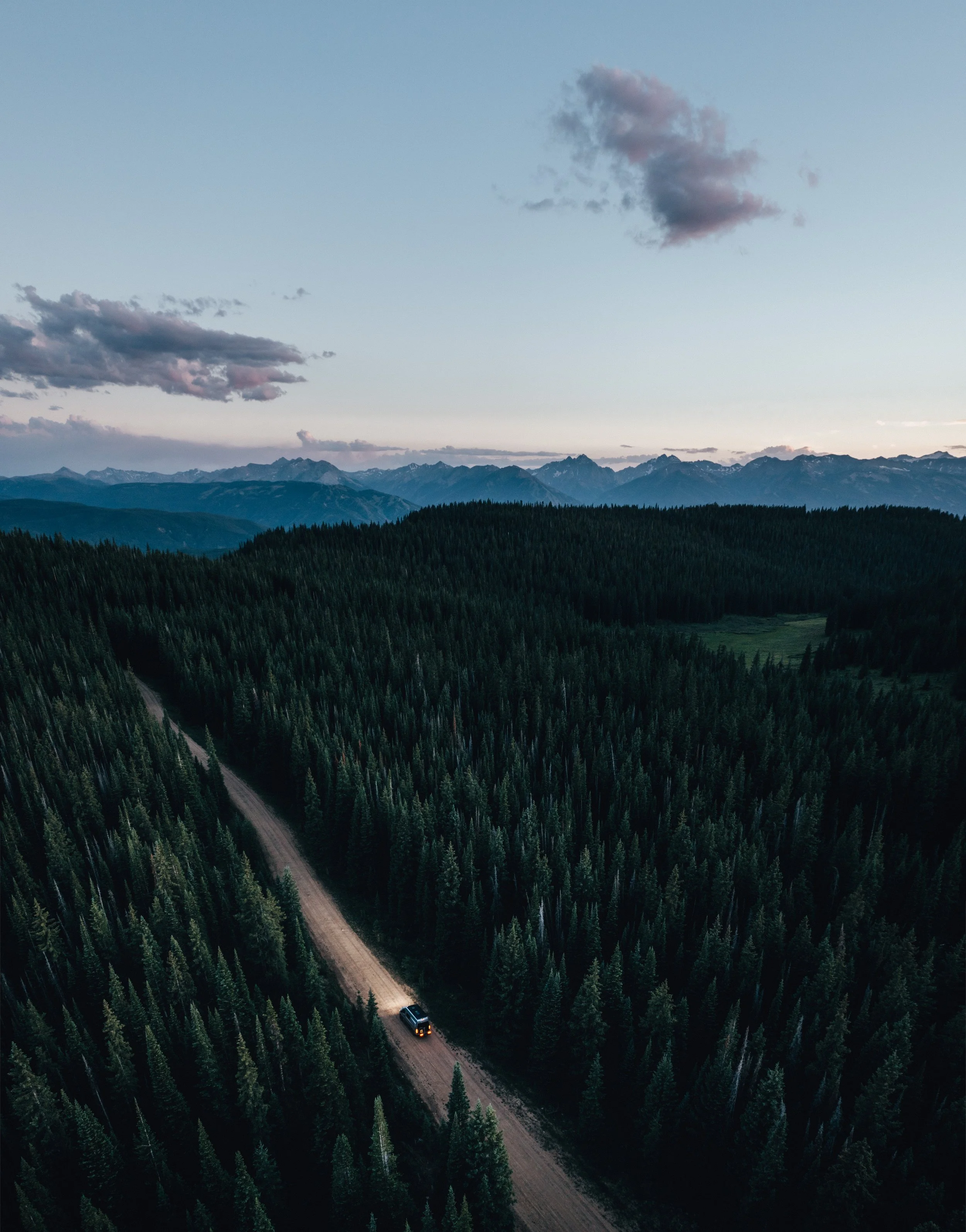 An aerial view of a forested mountain landscape at sunset with a dirt road running through the trees and a vehicle traveling on it.