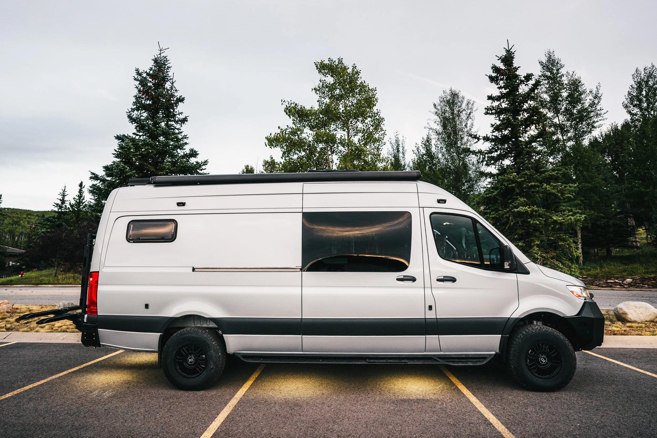 A silver camper van parked in a lot with yellow parking lines, surrounded by trees under an overcast sky.