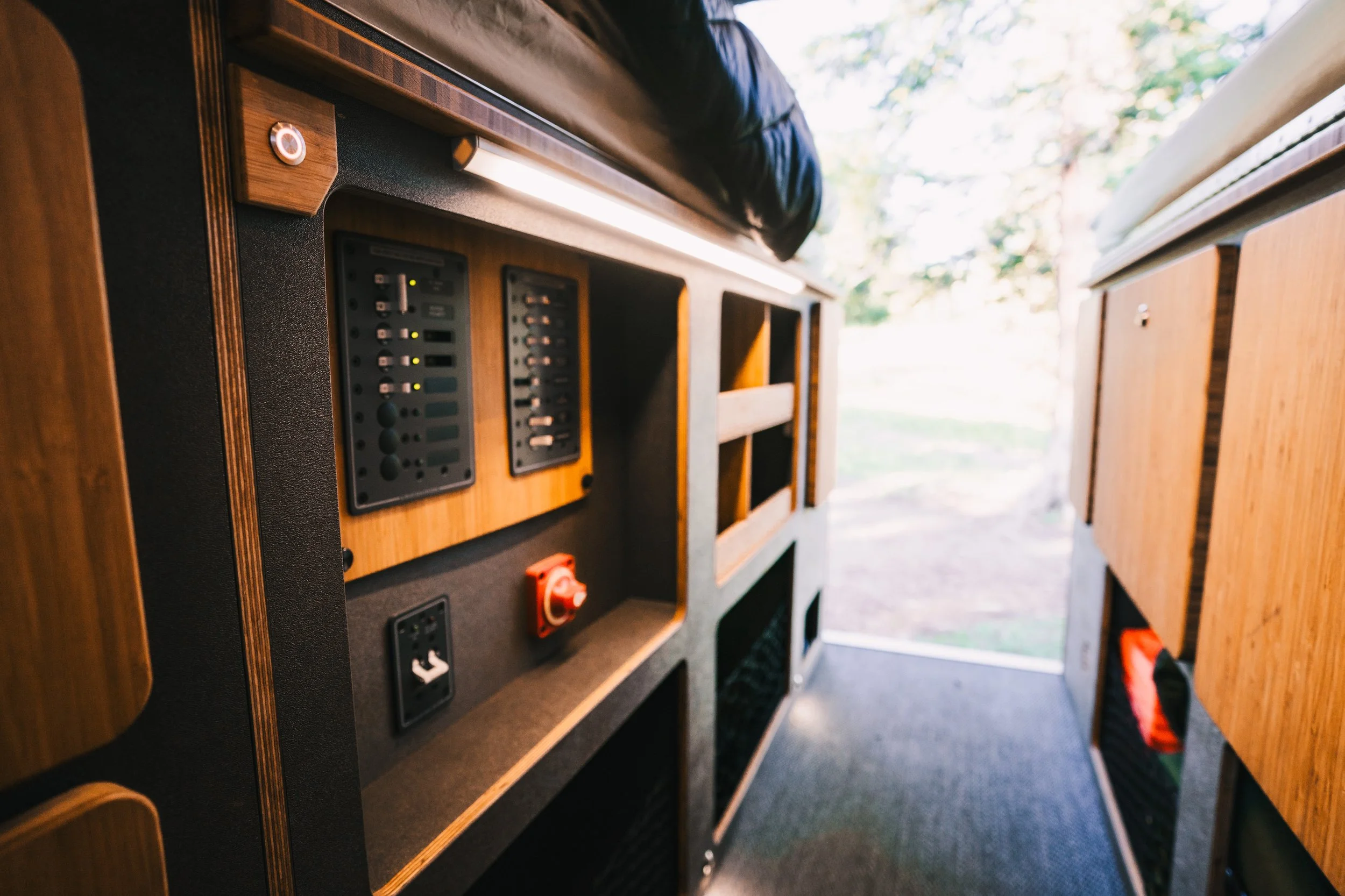 Interior of a wooden camper van with electrical outlets and controls on the side panel, and a view of trees outside.