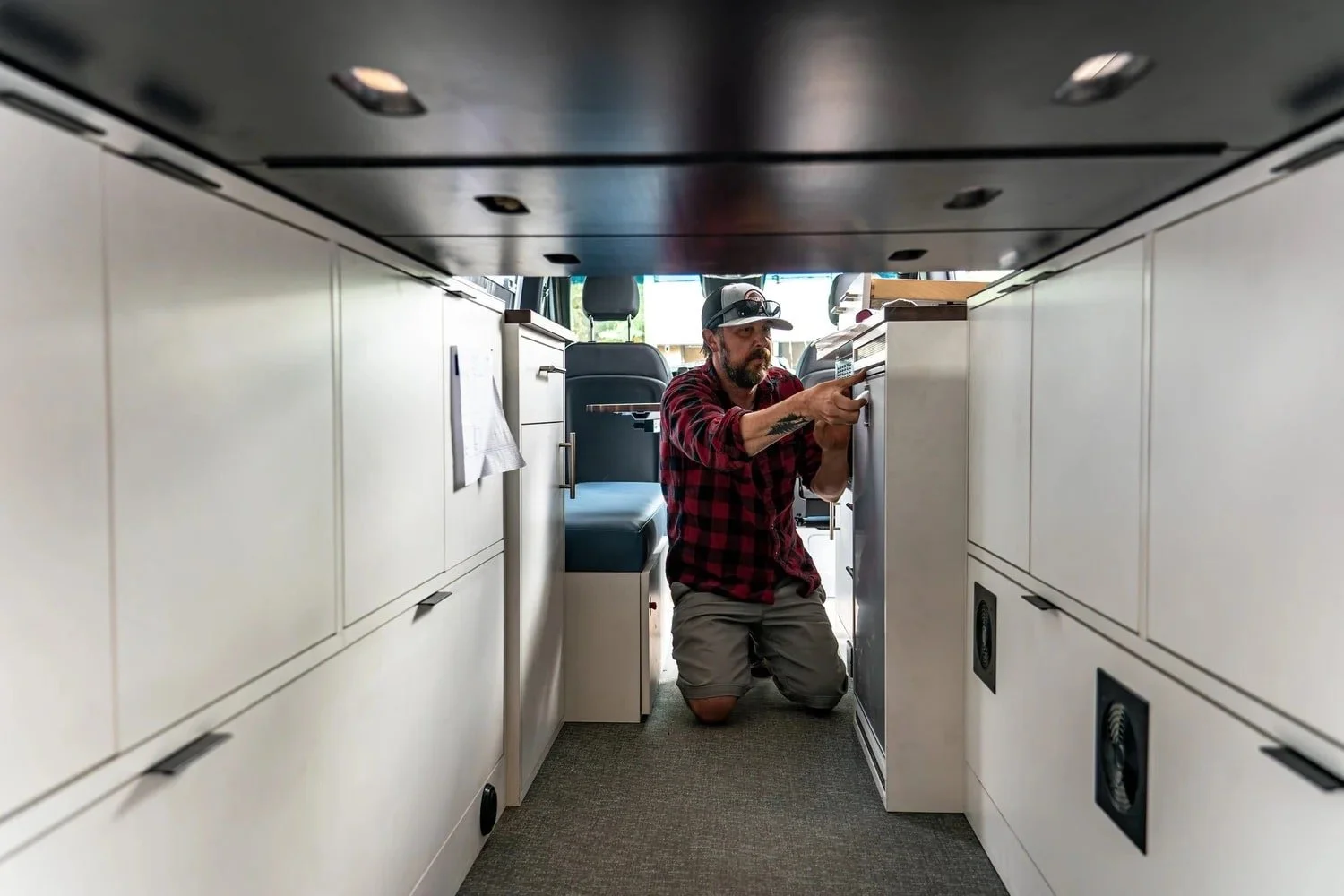 A man kneeling inside a small space between kitchen cabinets, working with a toolbox or drawer, wearing a red and black plaid shirt, grey shorts, a baseball cap, and sunglasses on his head.