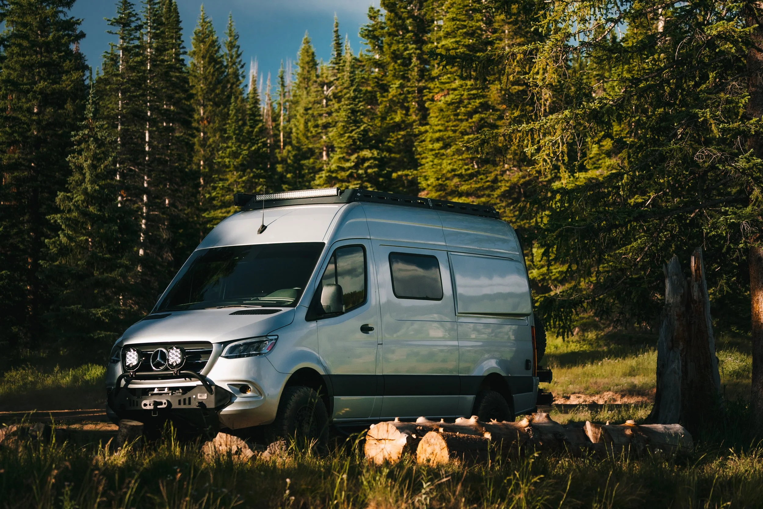 Silver camper van parked in a forest with tall pine trees and logs in the foreground.