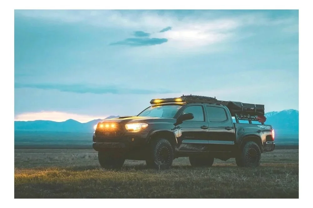 A black Toyota Tacoma pickup truck parked on a grassy field with mountains and a cloudy sky in the background during dusk or dawn.