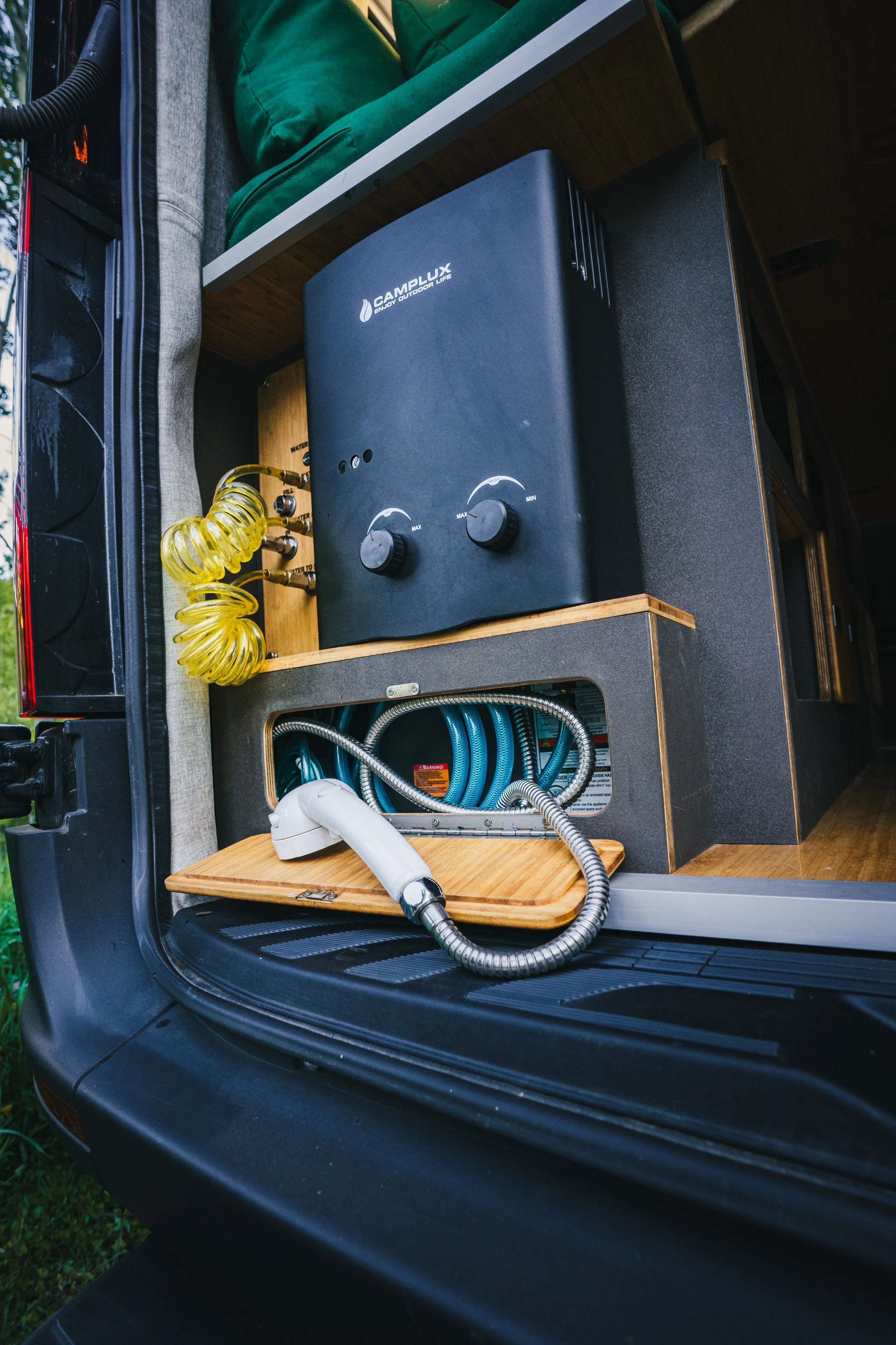 A close-up of a portable air compressor with yellow coiled air hoses, a black Camblux outdoor heater with control knobs, and blue hoses inside a wooden and fabric enclosure on the back of a vehicle.