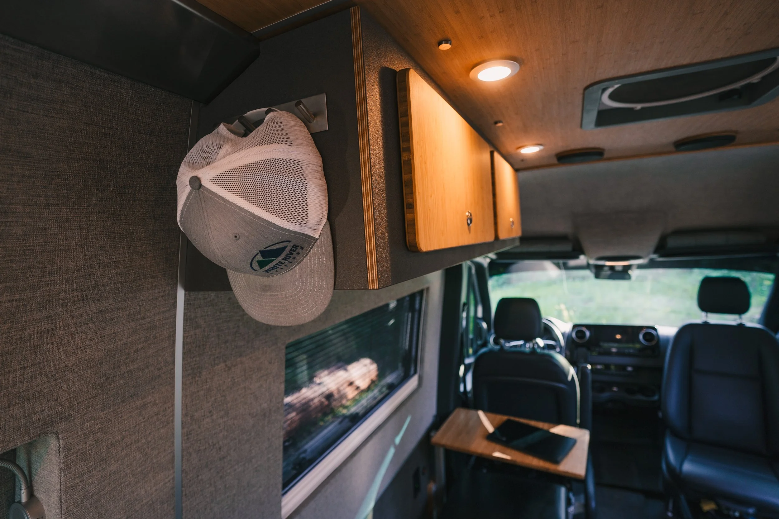 Interior of a vehicle, showing a baseball cap hanging on the wall, a small wooden cabinet with lighting, and part of the driver's area with seats and a window.