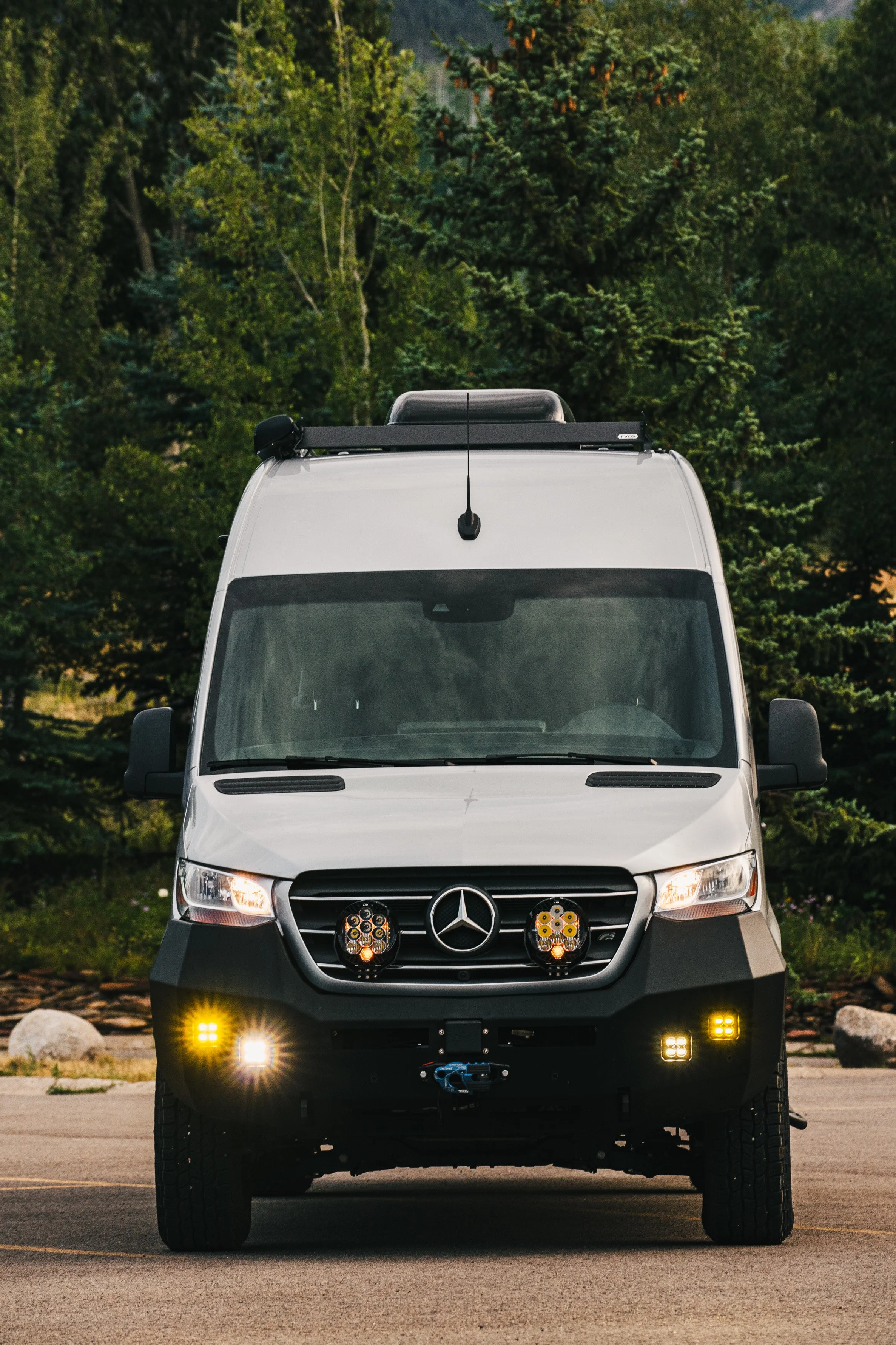 Front view of a silver Mercedes-Benz Sprinter van equipped with off-road lights, camping gear on the roof, and a front tow hook, parked on pavement with trees in the background.