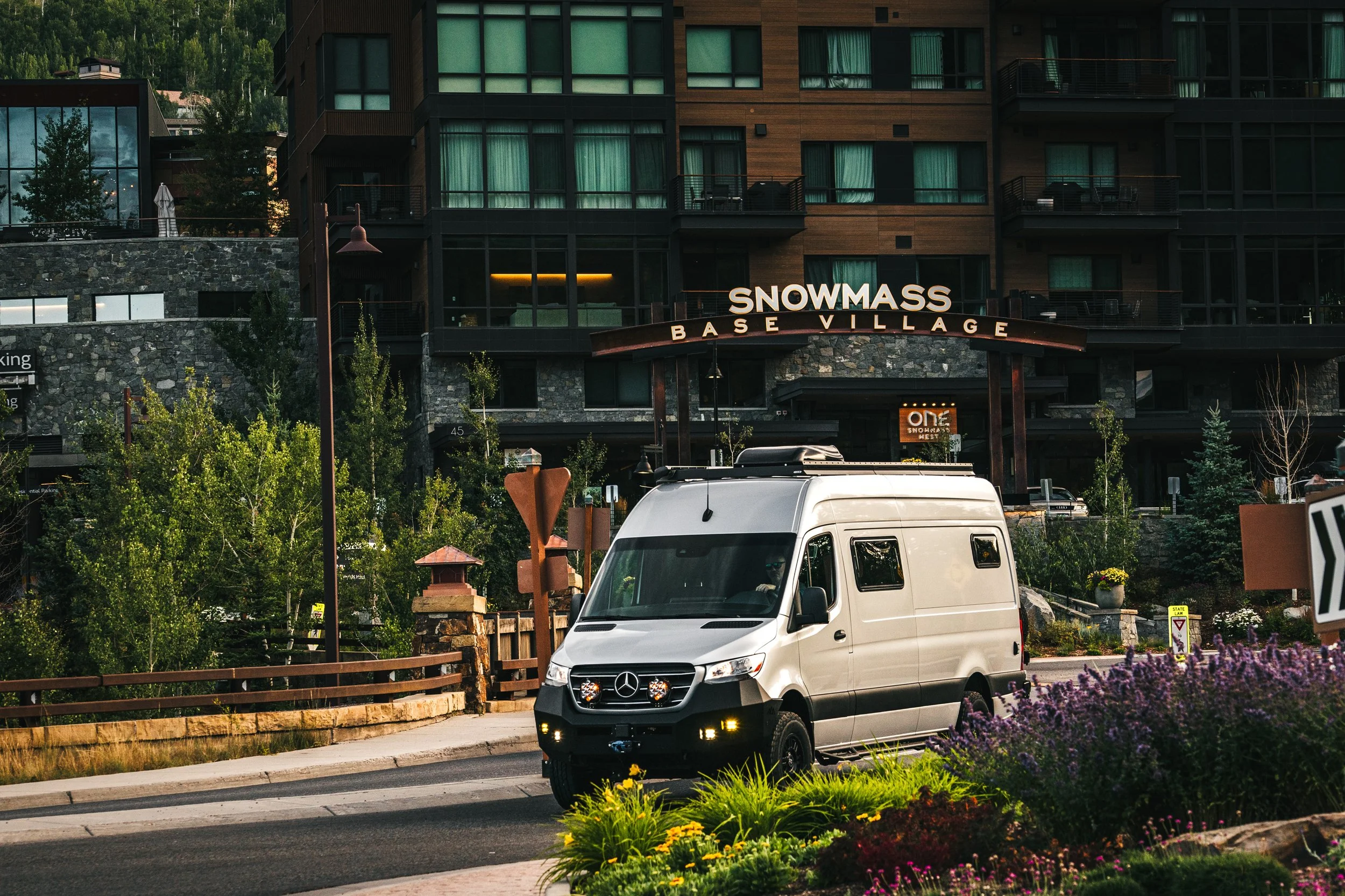 A silver Mercedes-Benz Sprinter van driving on a road in front of a building with a sign that says 'Snowmass Base Village', surrounded by trees and flowers.