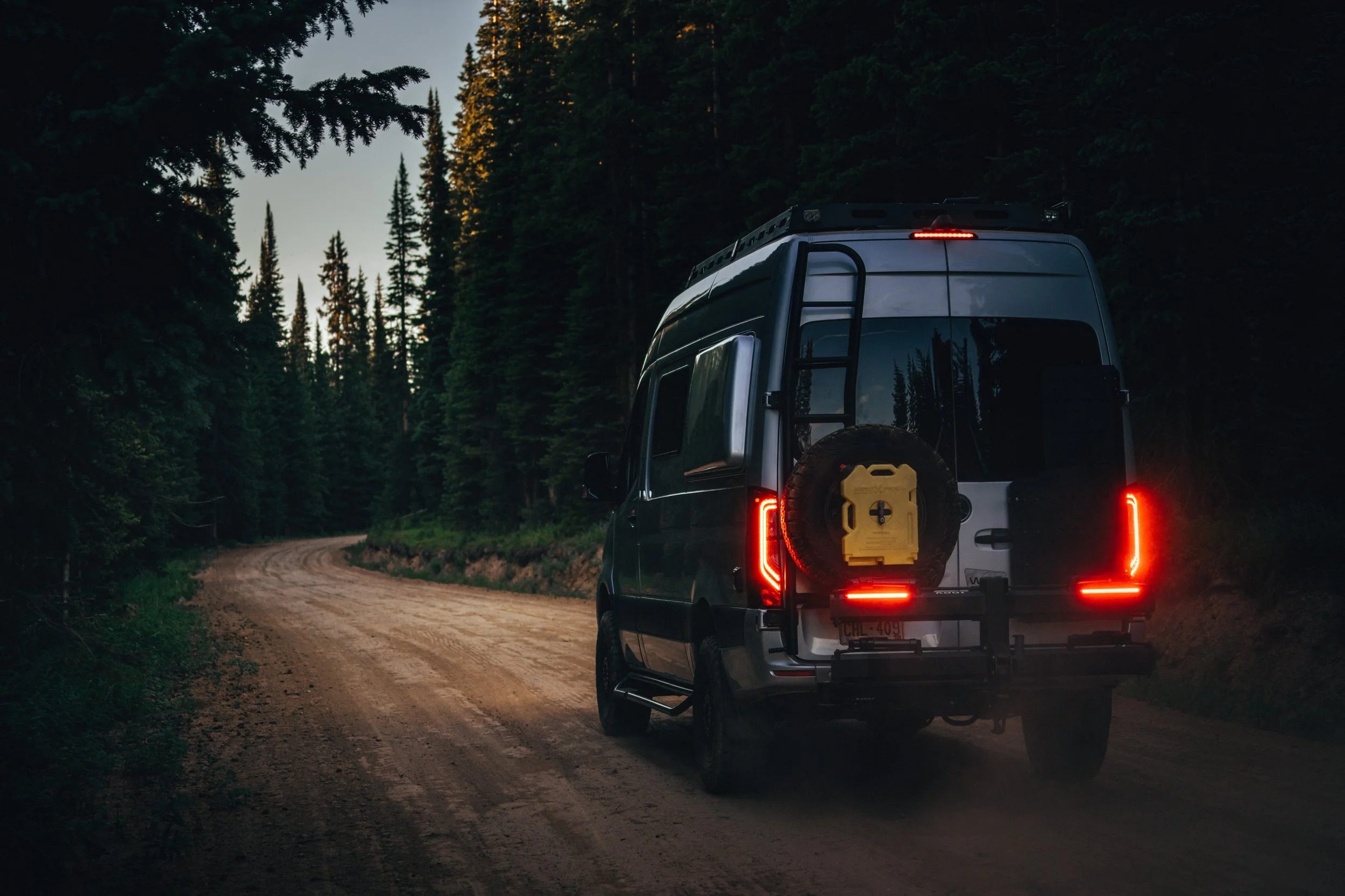 A dark-colored van driving on a winding dirt road through a dense forest of tall evergreen trees during dusk.