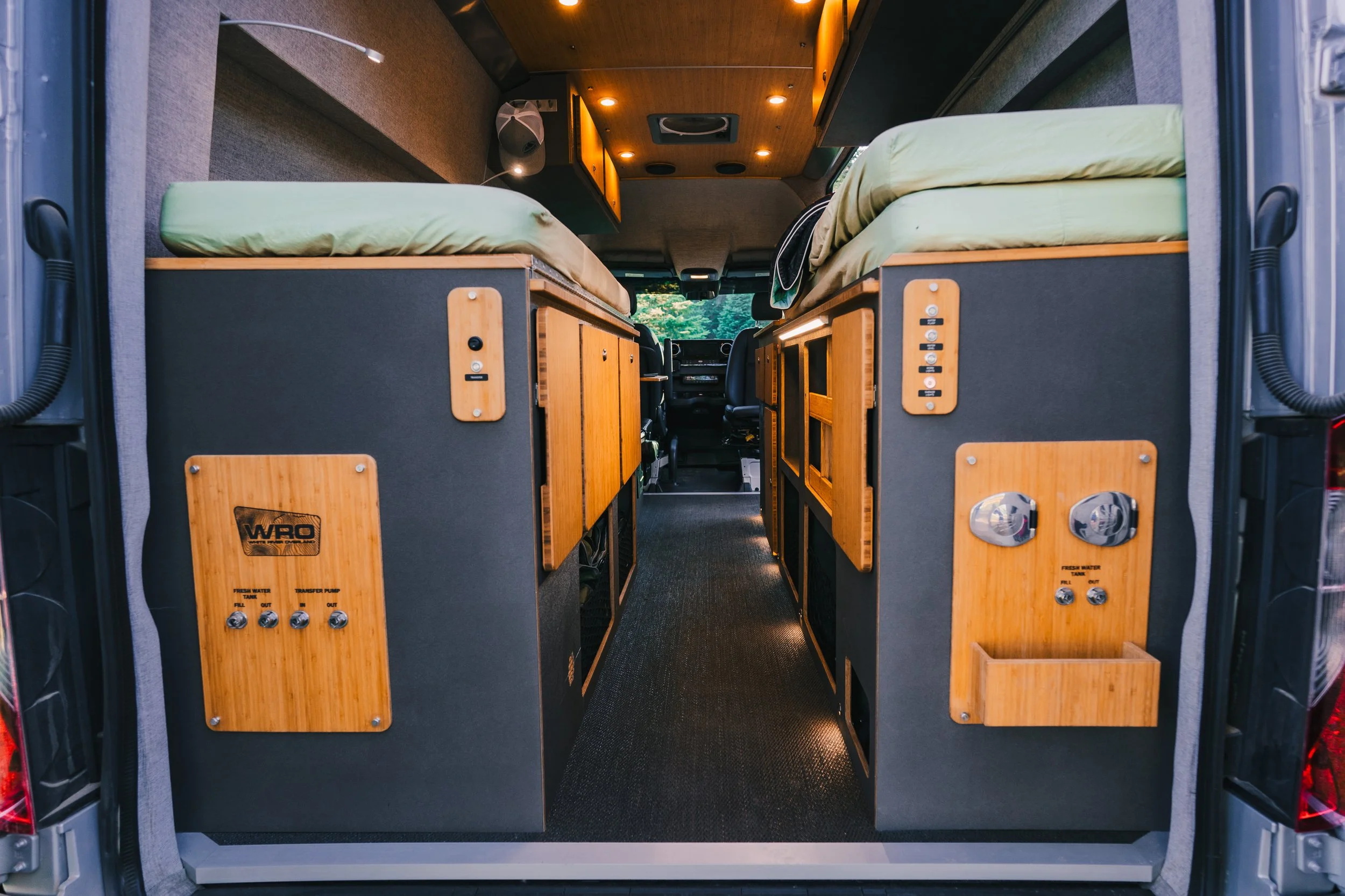 Interior of a camper van showing two beds on either side, with wooden cabinetry, control panels, and a corridor in the middle, with the driver and passenger seats visible in the background.