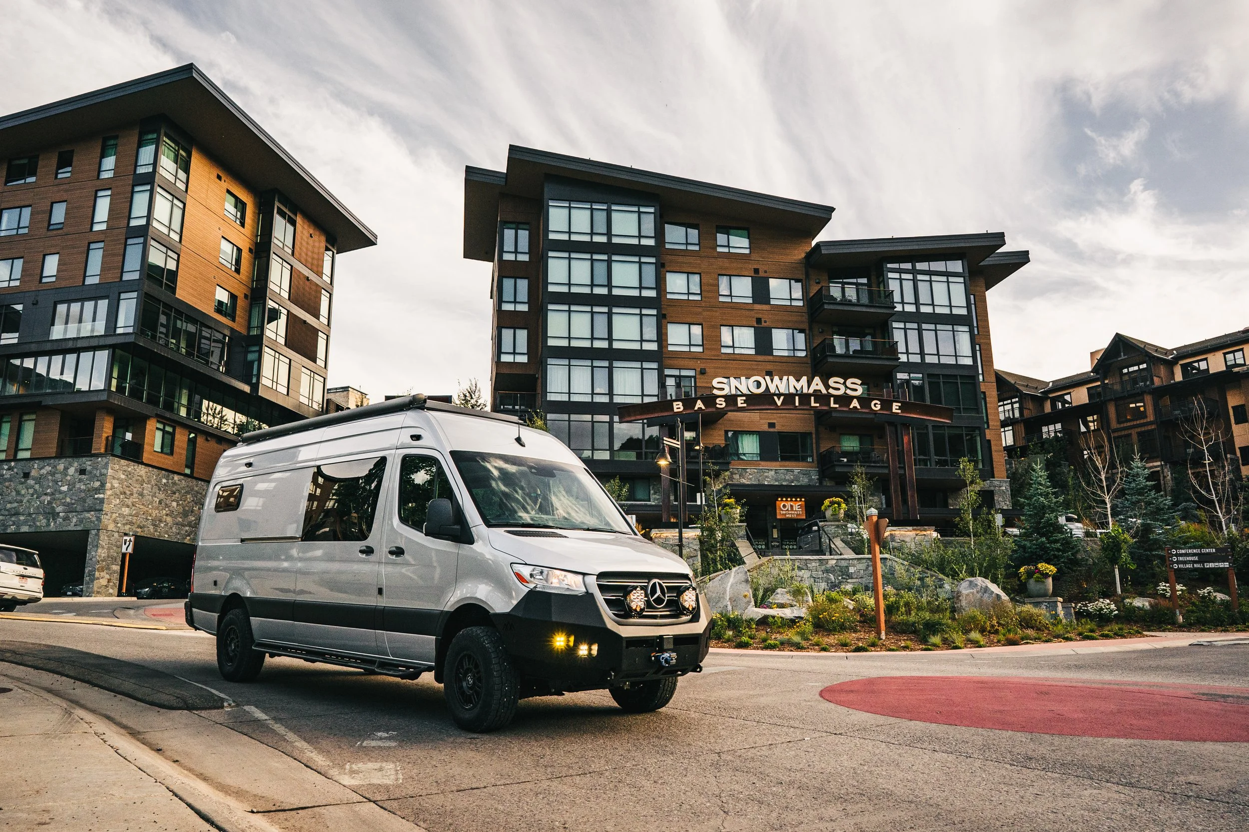 Modern multi-story apartment building with sign reading 'Snowmass Base Village' and a parked Silver Mercedes-Benz van in the foreground.