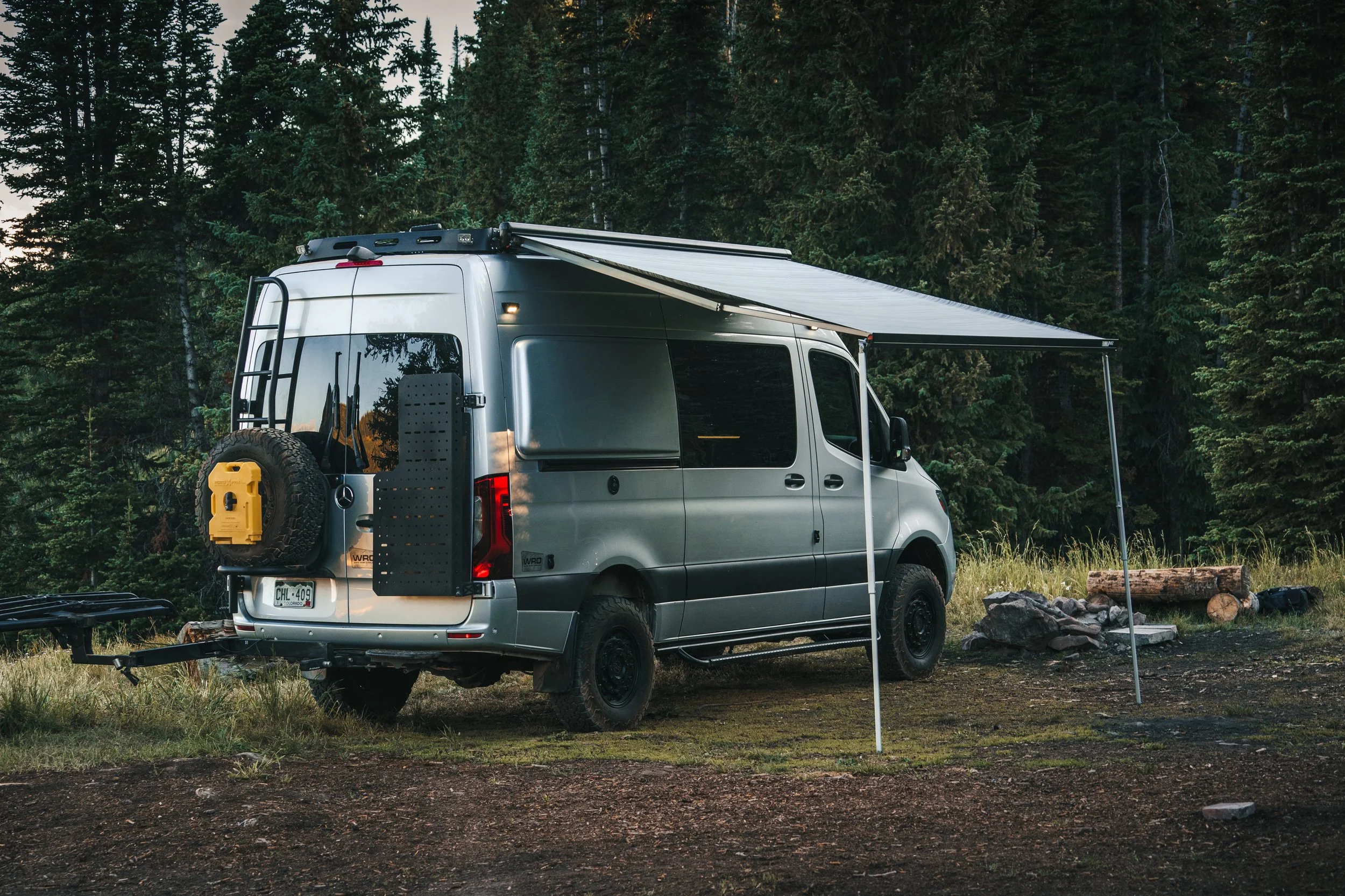 Silver camper van with extended awning parked outdoors in a forested area with logs and rocks on the ground.