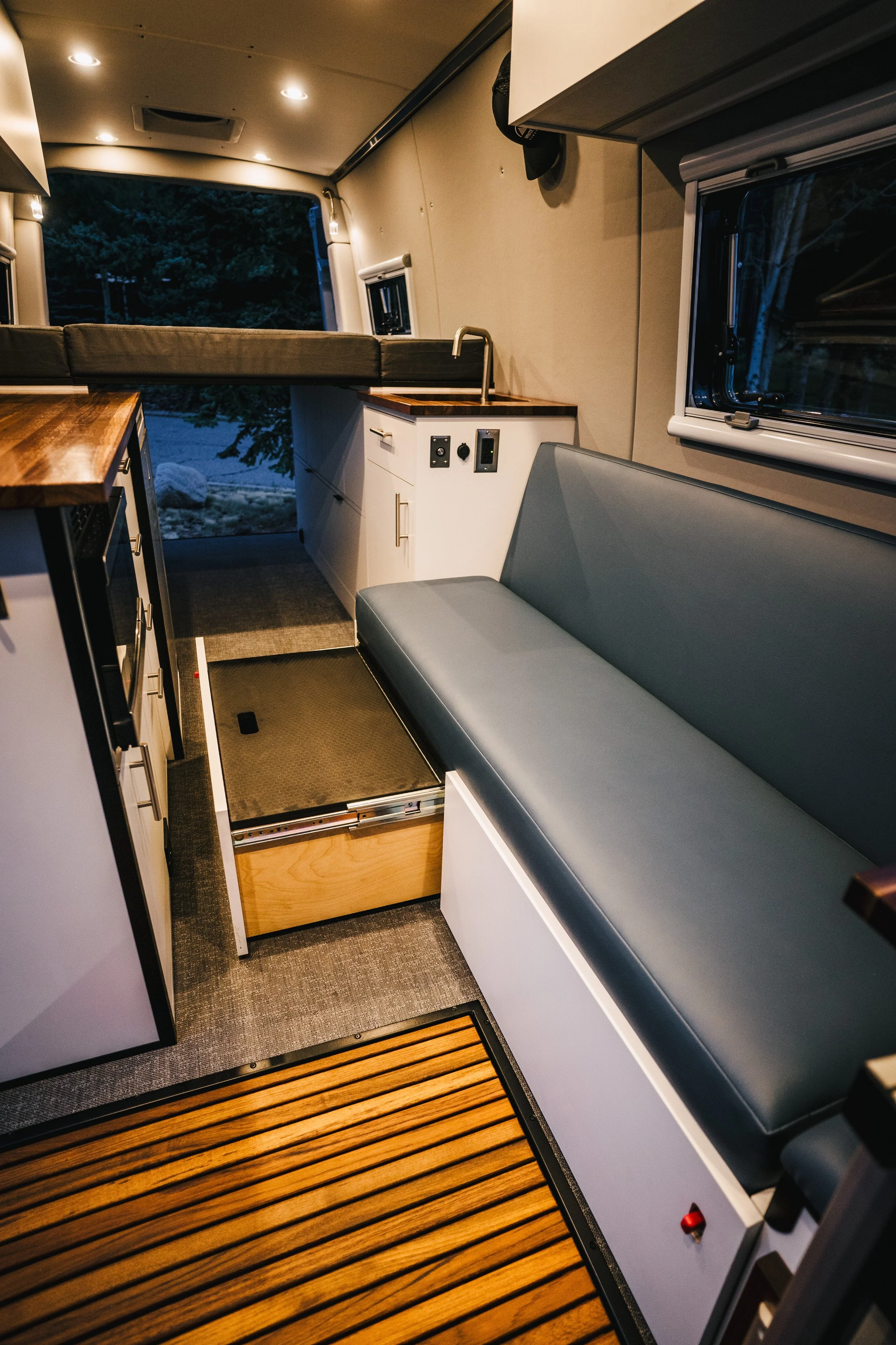 Interior of a camper van showing a bench seat, a small kitchenette with a sink and countertop, and a view outside through a window.