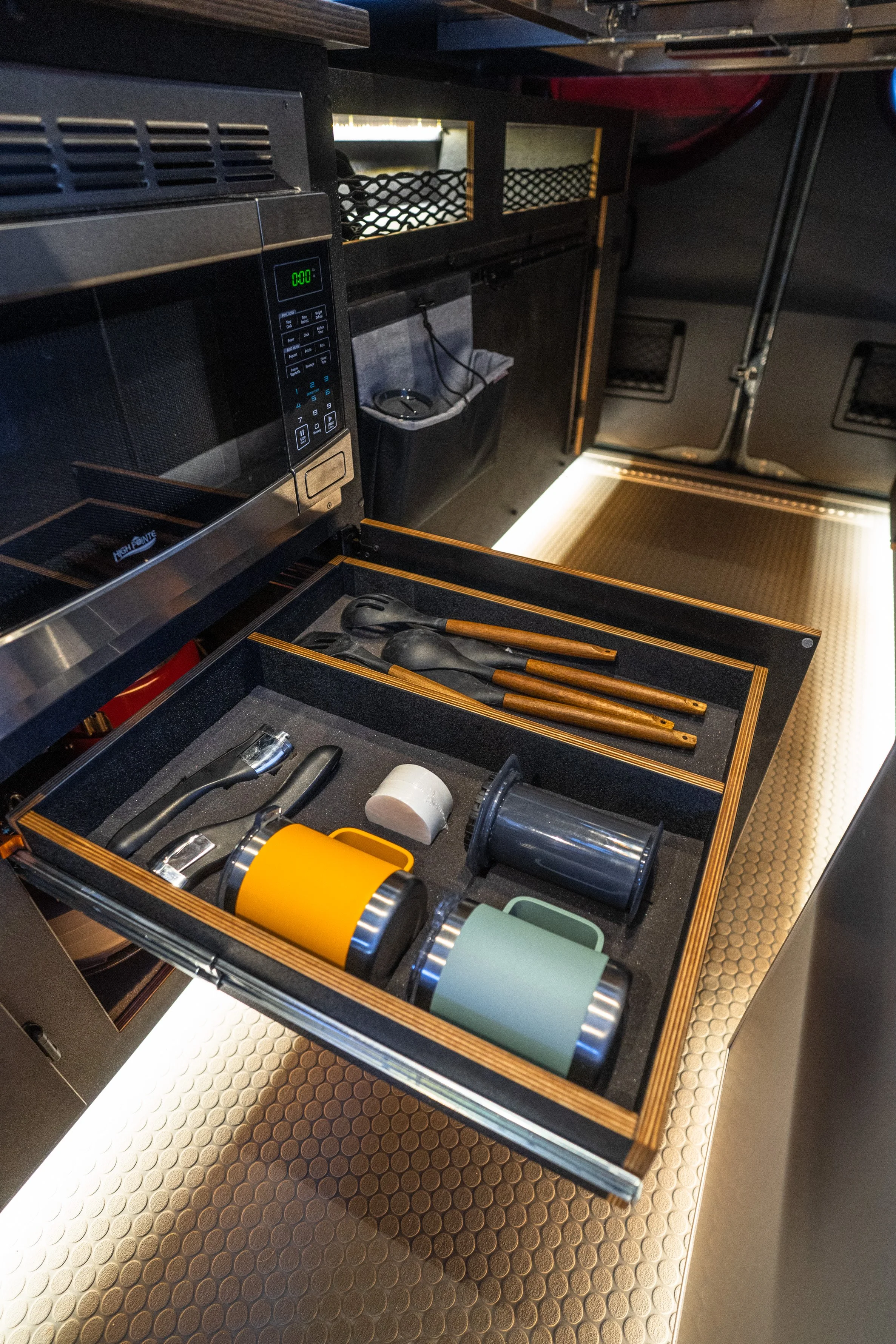 Open kitchen drawer with utensils, cups, and a roll of tape inside a kitchen with stainless steel appliances.