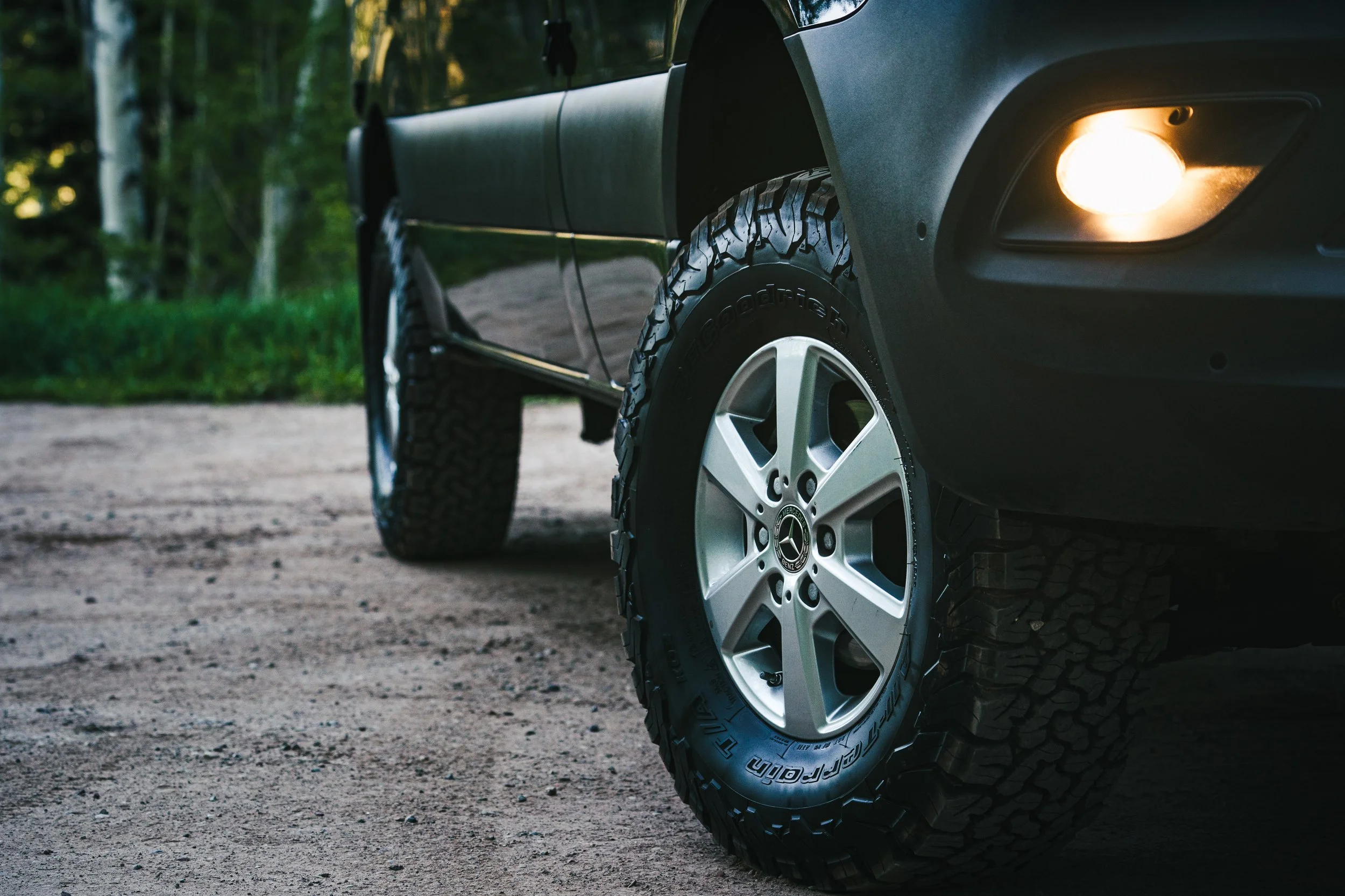 Close-up of a black SUV's front side with a lit fog light, large off-road tire, and silver alloy wheel, parked on a dirt trail surrounded by trees.