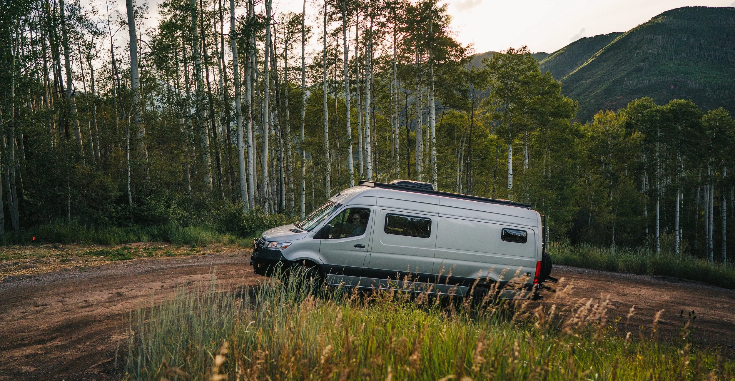 A silver camper van driving on a dirt road surrounded by green grass and trees, with mountains in the background at sunset.