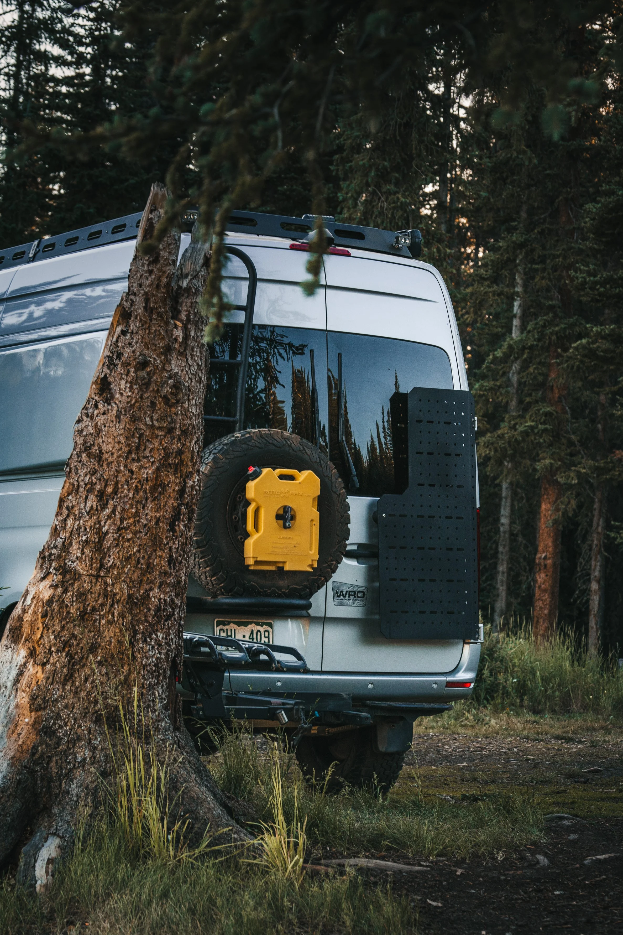 Rear view of a silver van parked in a forested area, showing a mounted spare tire with a yellow jerry can attached, a black pegboard, and a license plate reading 'SHU 409'.