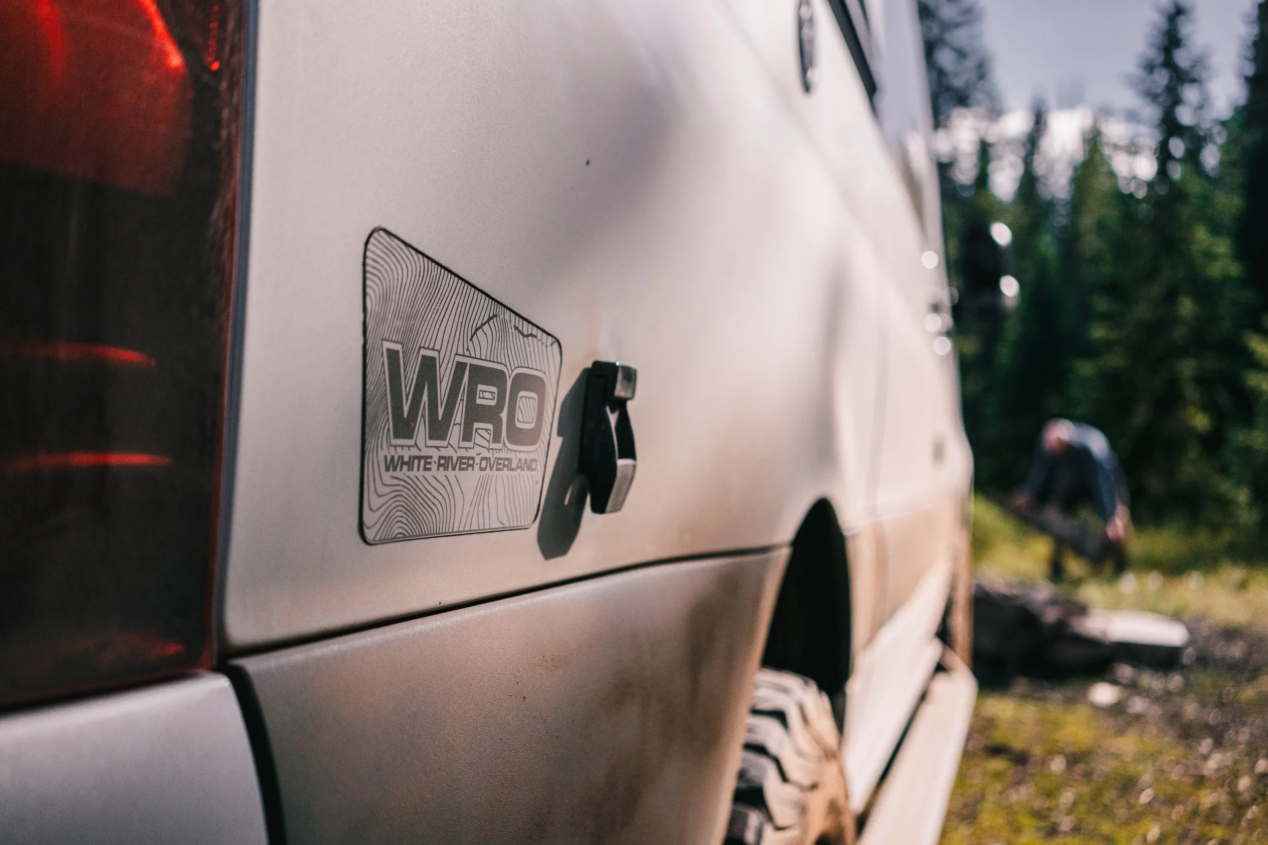Close-up of a beige vehicle with a 'White River Overland' (WRO) sticker, showing part of the rear and side, tire, and a person in the background in a wooded outdoor setting.