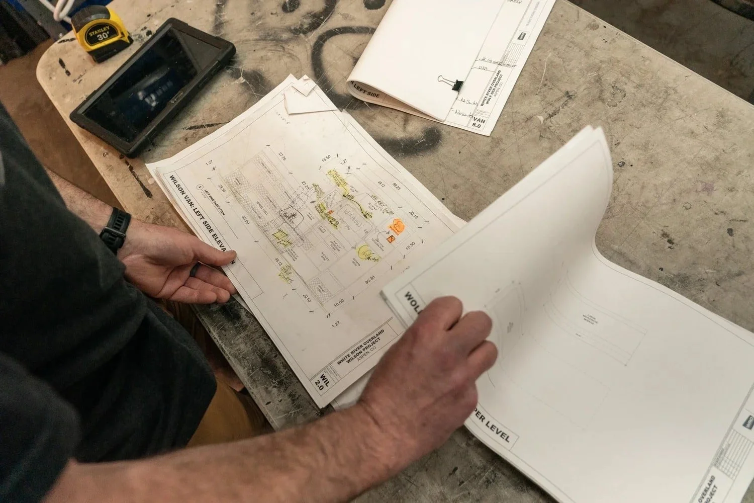 A person reviewing van build blueprints on a work table with a calculator, a smartphone, and additional papers.