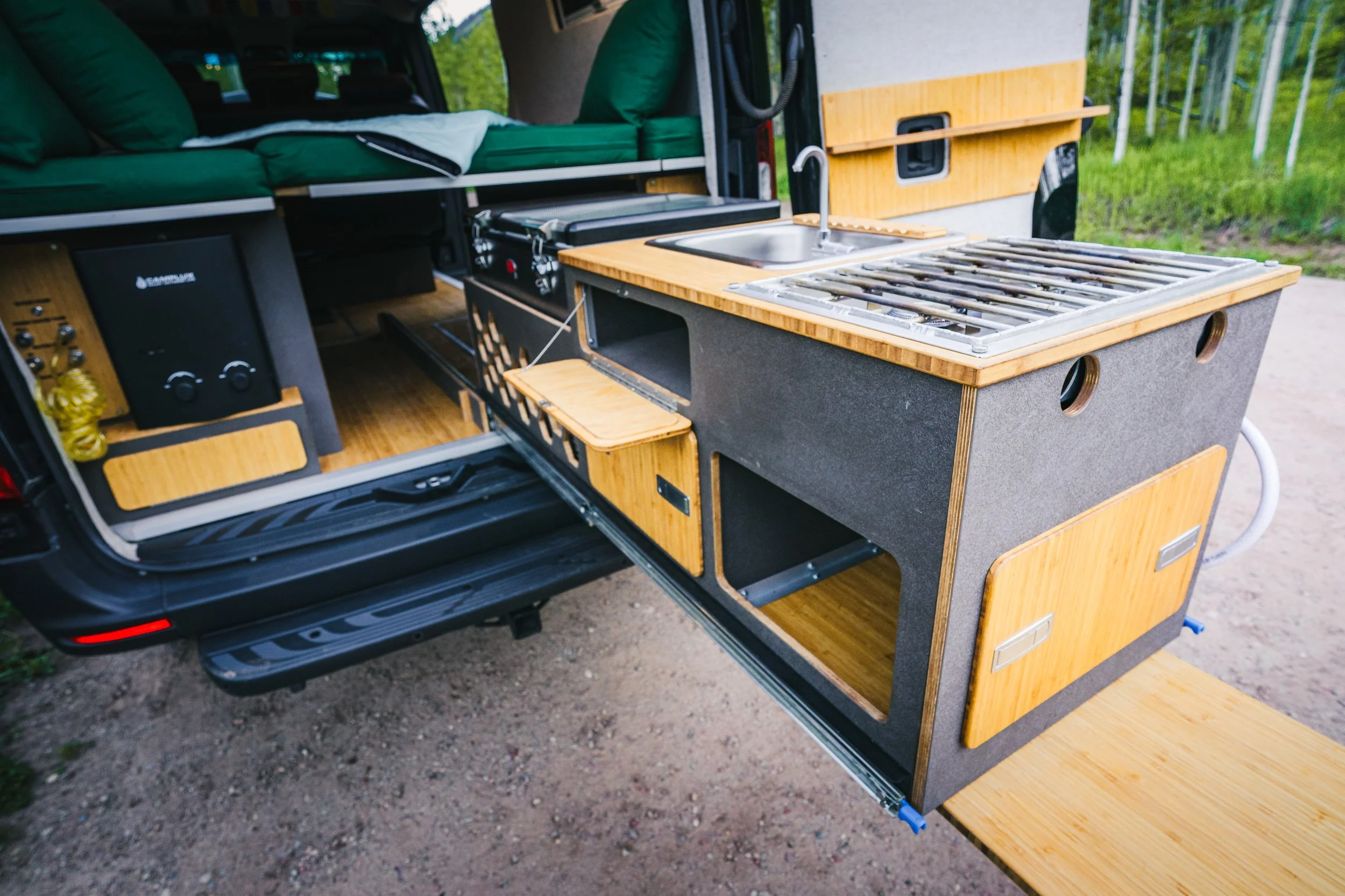 Camper van interior with wooden kitchen cabinets, a stovetop, sink, and seating area with green cushions, set in a forested area.