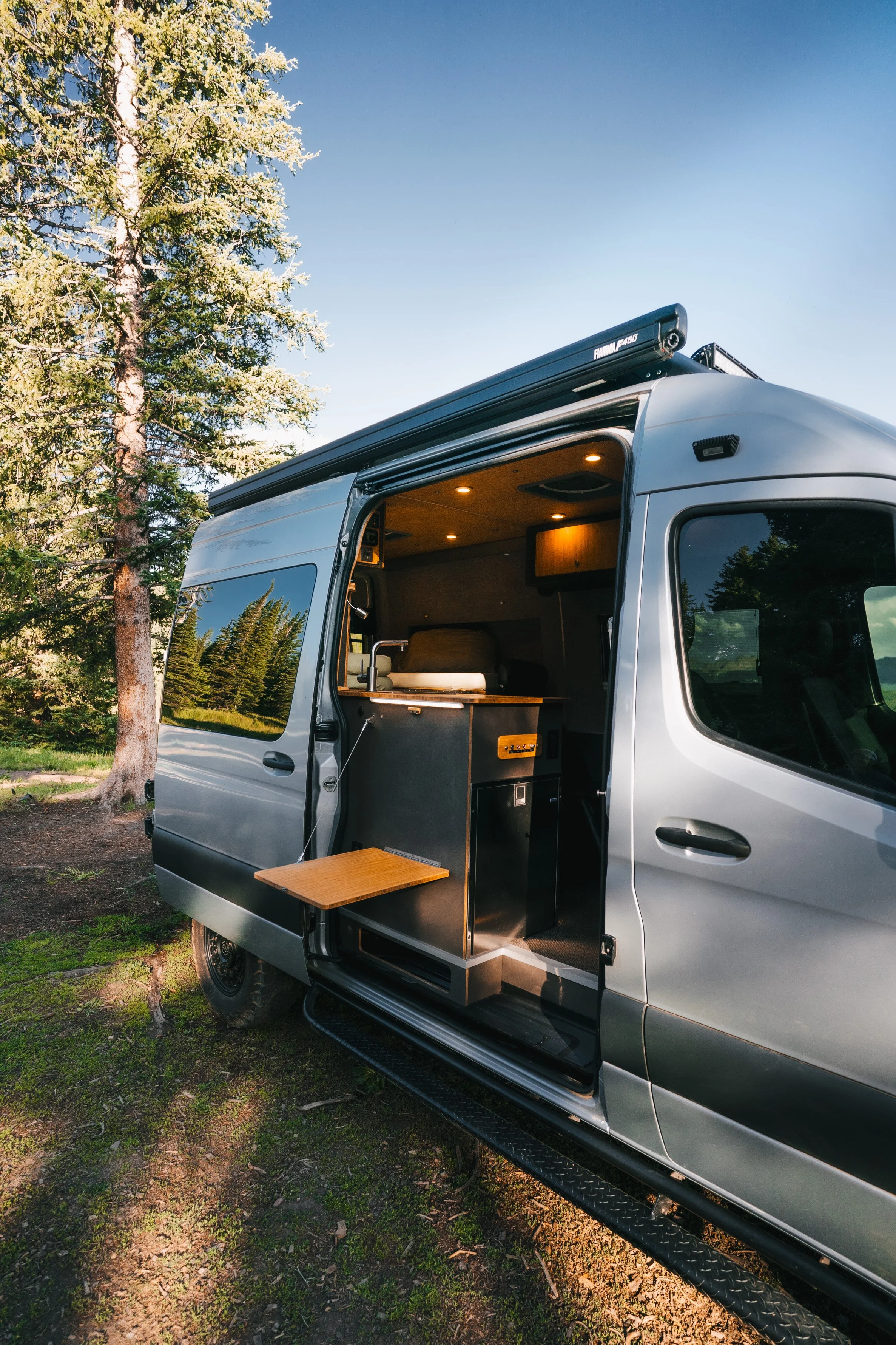 A silver camper van parked in a wooded area, with its slide-out section extended, showing a small kitchen with a countertop and wood-paneled ceiling.