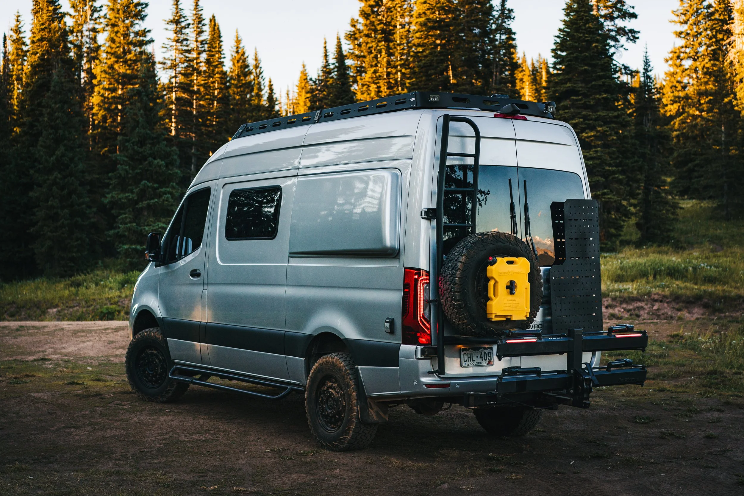 Silver camper van parked outdoors on dirt with a forested background during sunset. The back of the van has an external spare tire, a yellow toolbox, and a black luggage rack.