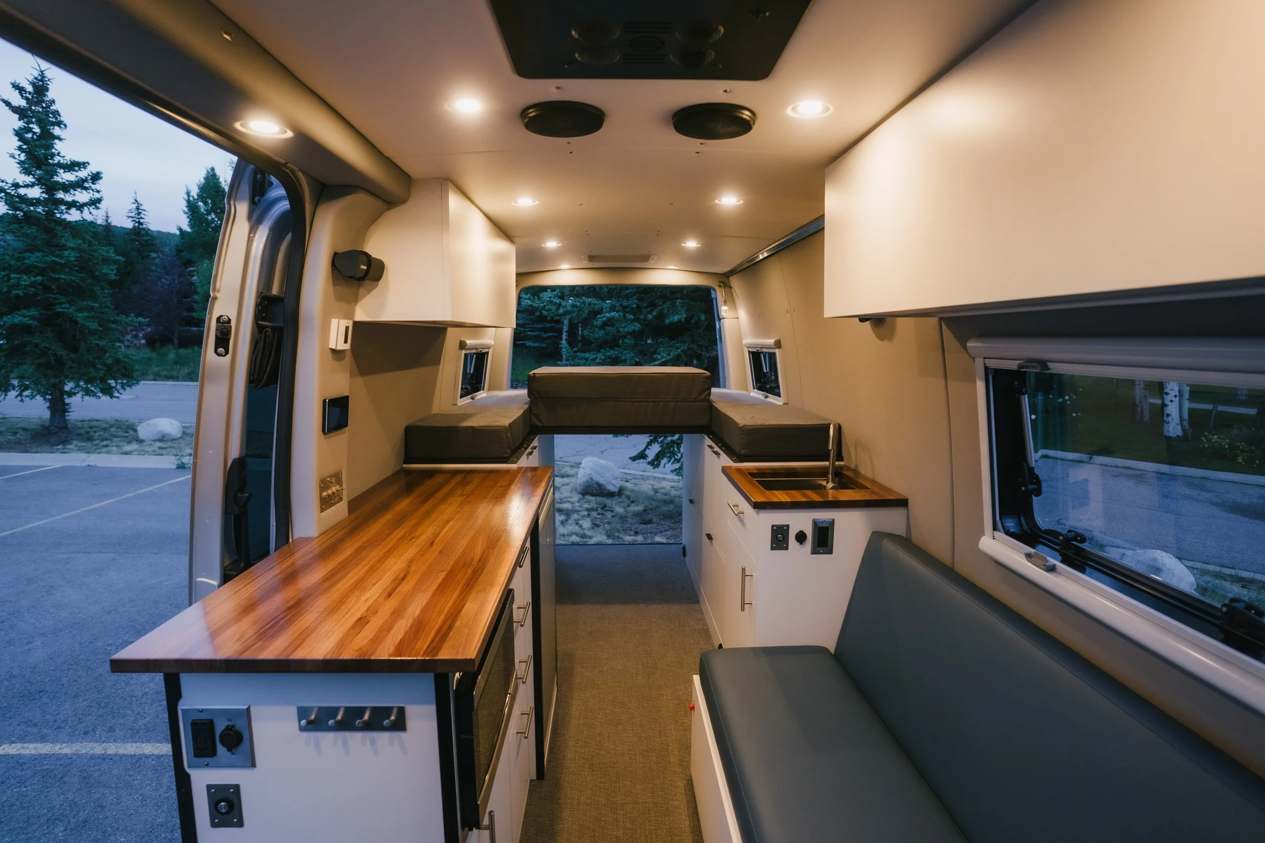 Interior of a converted van with a wooden countertop, windows, cushioned seating, and a small kitchen area.