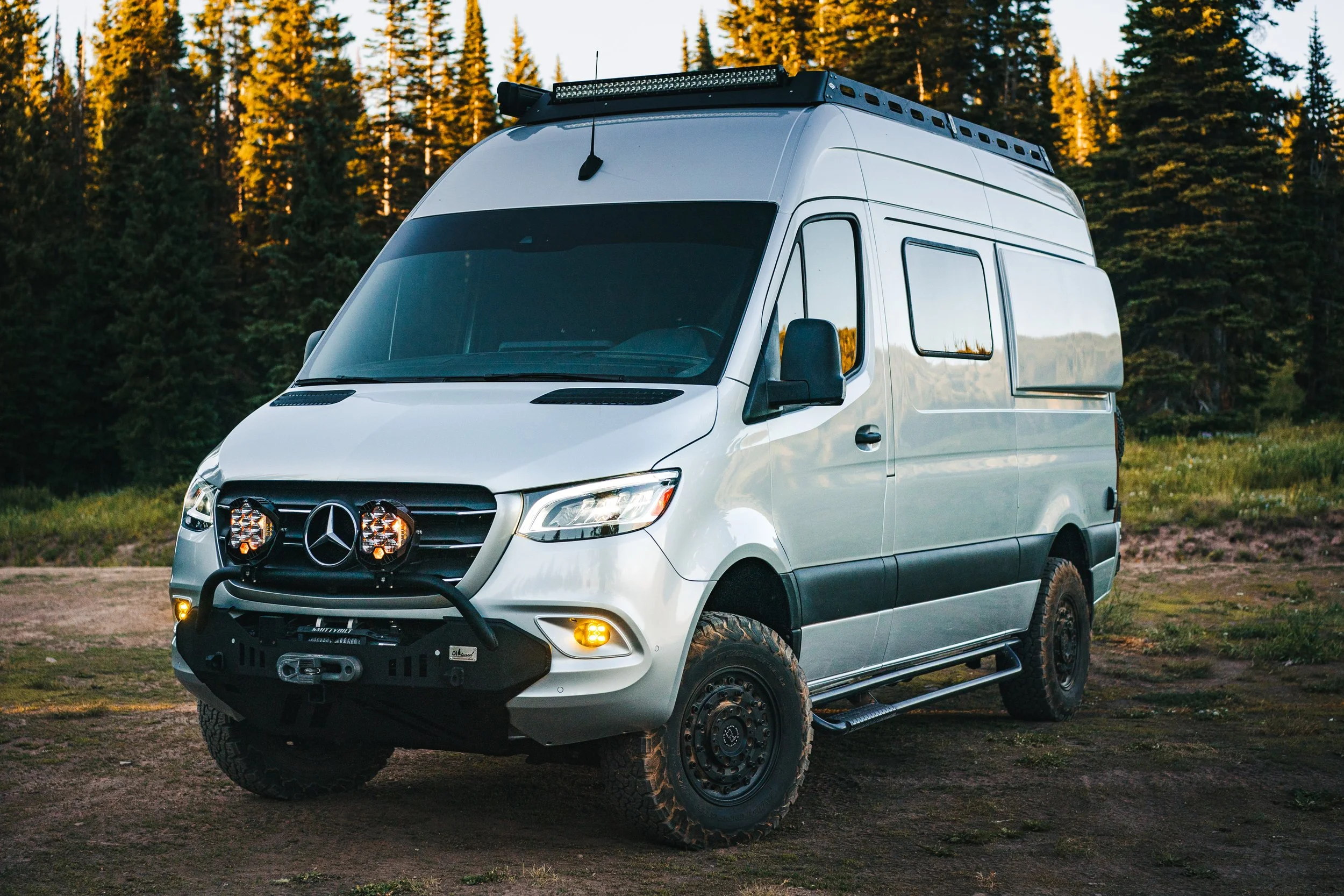 A white Mercedes-Benz Sprinter van off-road in a forested area during daylight.