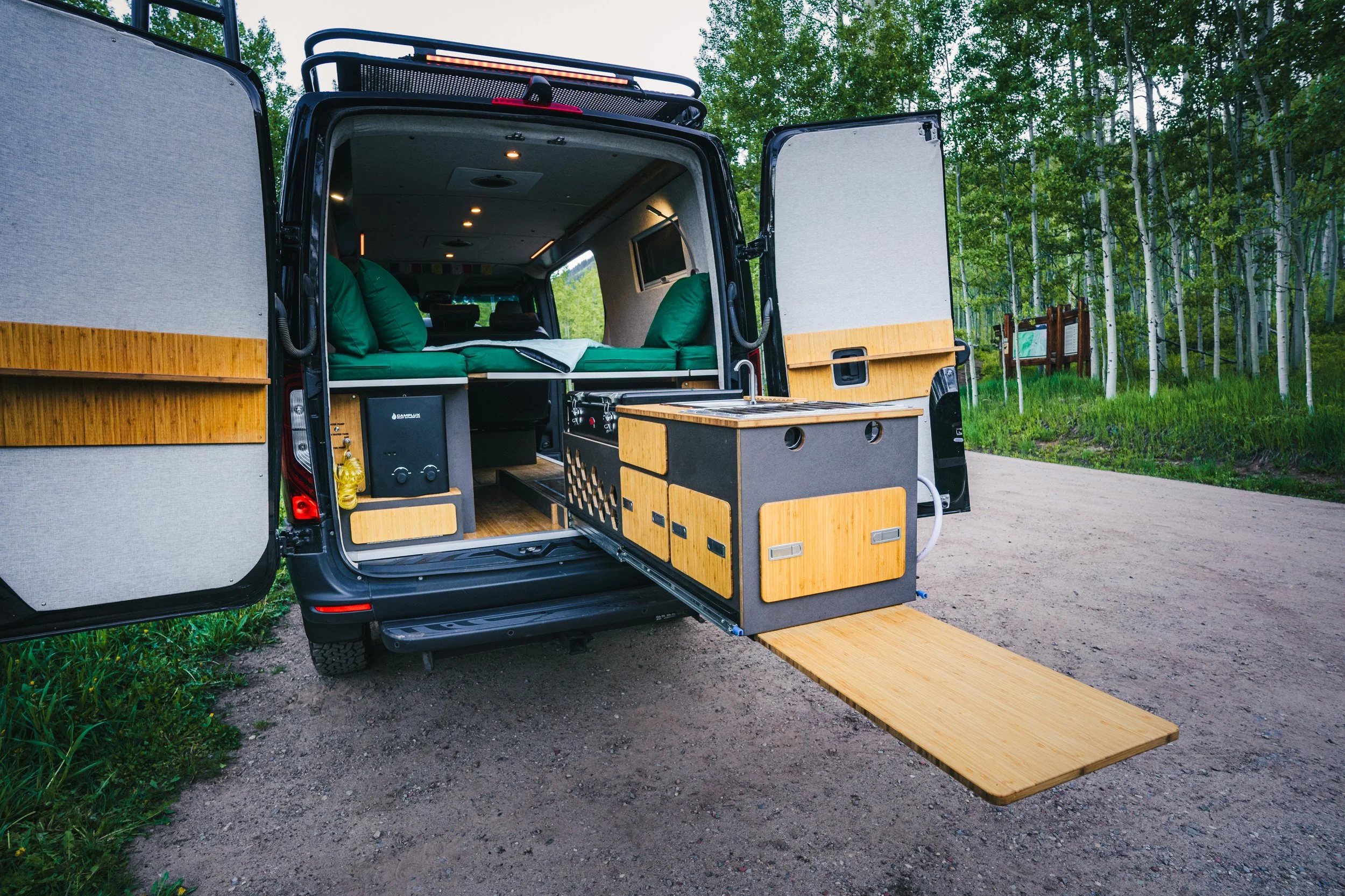 Interior of a black camper van with custom wooden storage and a fold-out wooden deck, parked in a forested area.