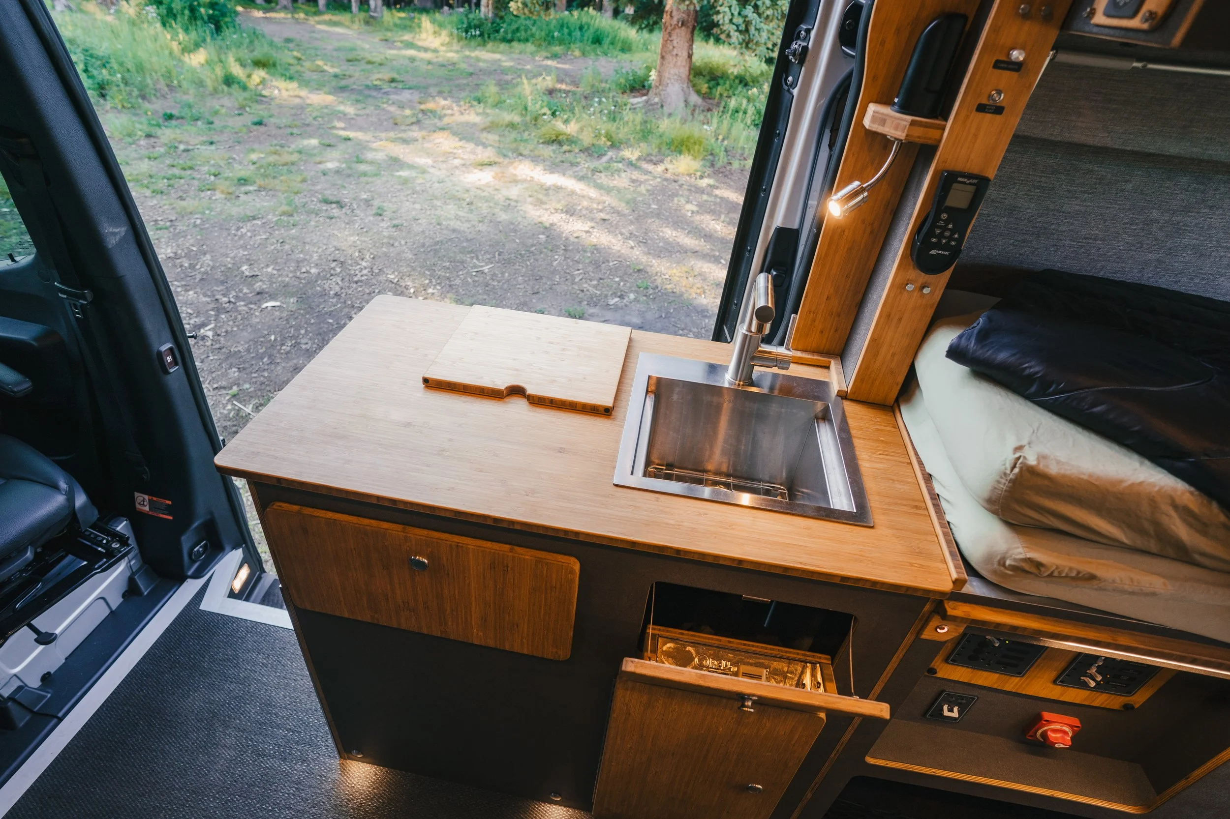 Interior of a camper van with a small wooden kitchen area, including a metal sink with a faucet, a cutting board, a bed with a black pillow, and a view of trees and grass outside through the open door.