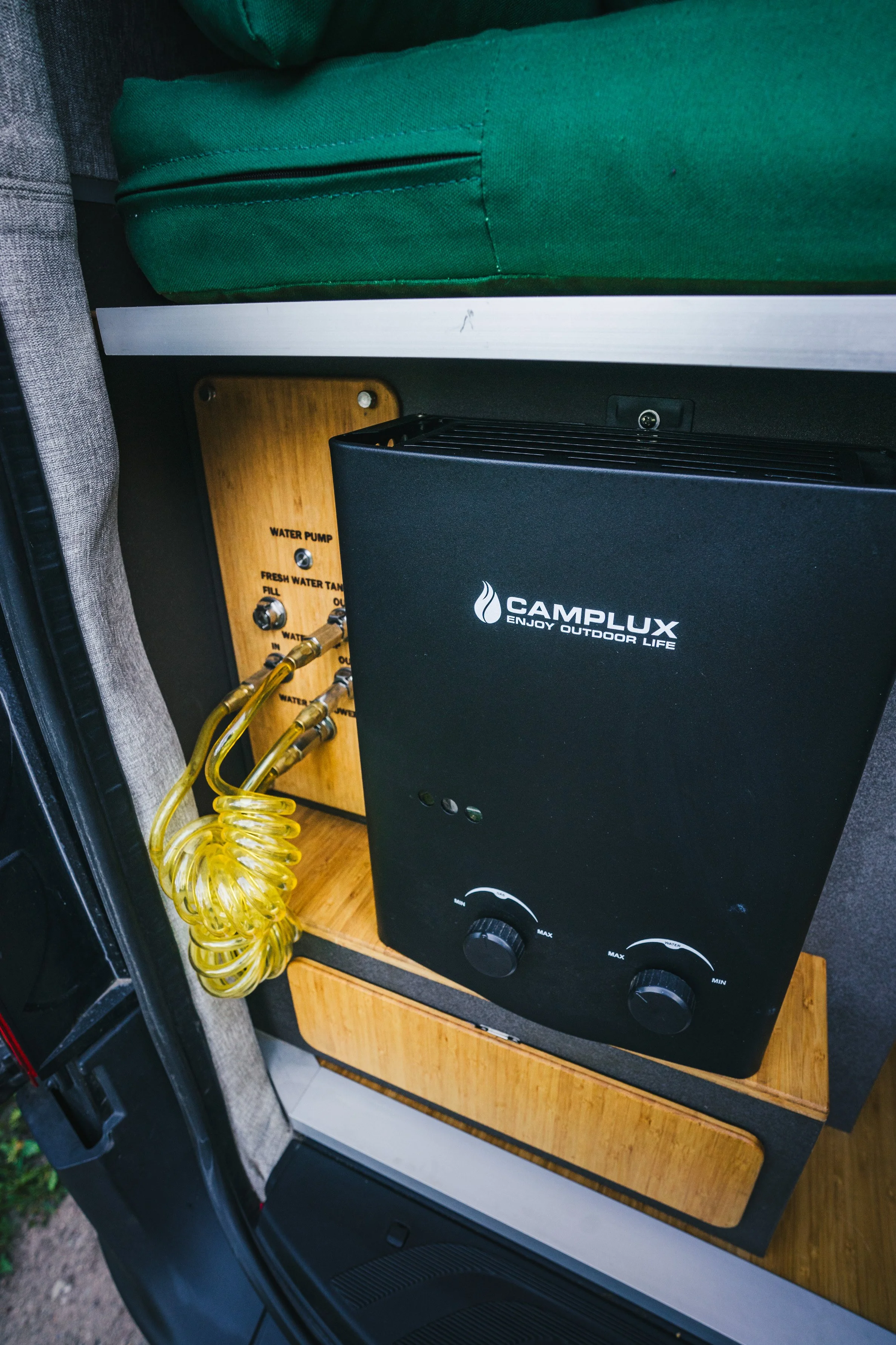 A black portable water heater labeled 'CAMP.LUX ENJOY OUTDOOR LIFE' placed on a wooden shelf inside a vehicle. Behind it, there is a wooden panel with water pump and water tank gauges, and yellow coiled water hoses.