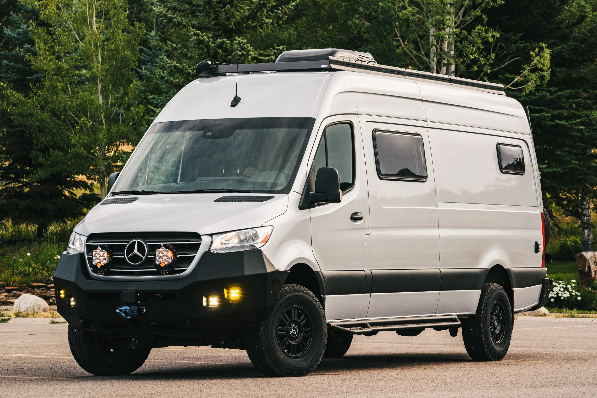 Silver Mercedes-Benz Sprinter van parked on the side of a road with green trees in the background.