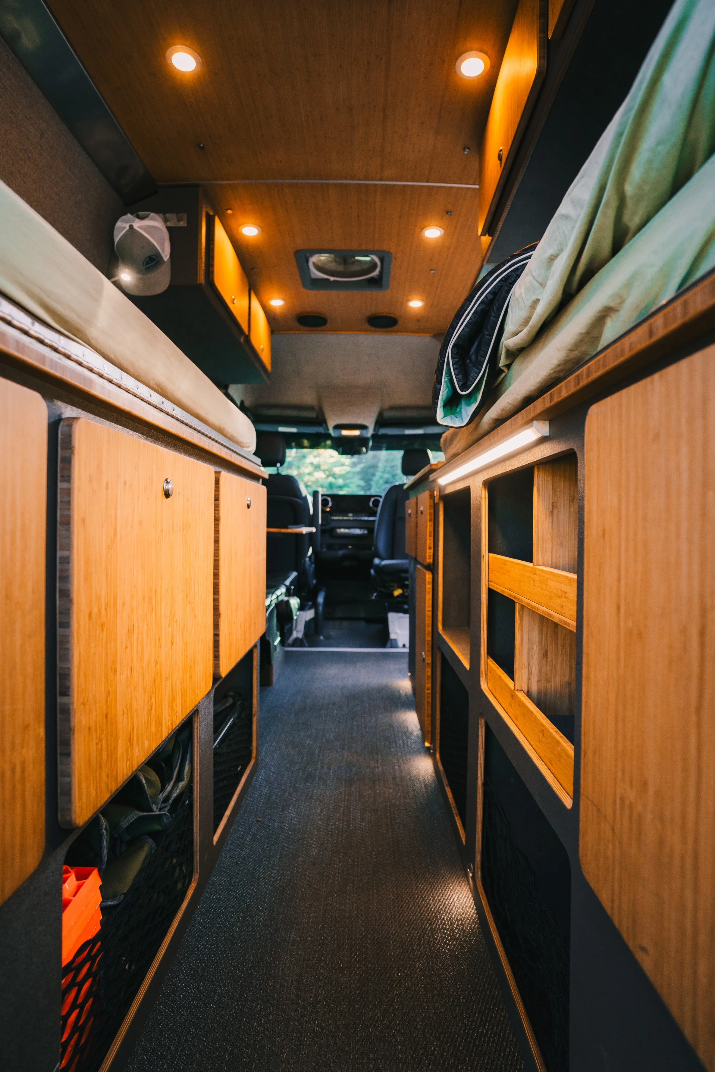 Interior view of a camper van with wooden cabinets and a narrow aisle between sleeping areas on either side, leading to the driver's cab.