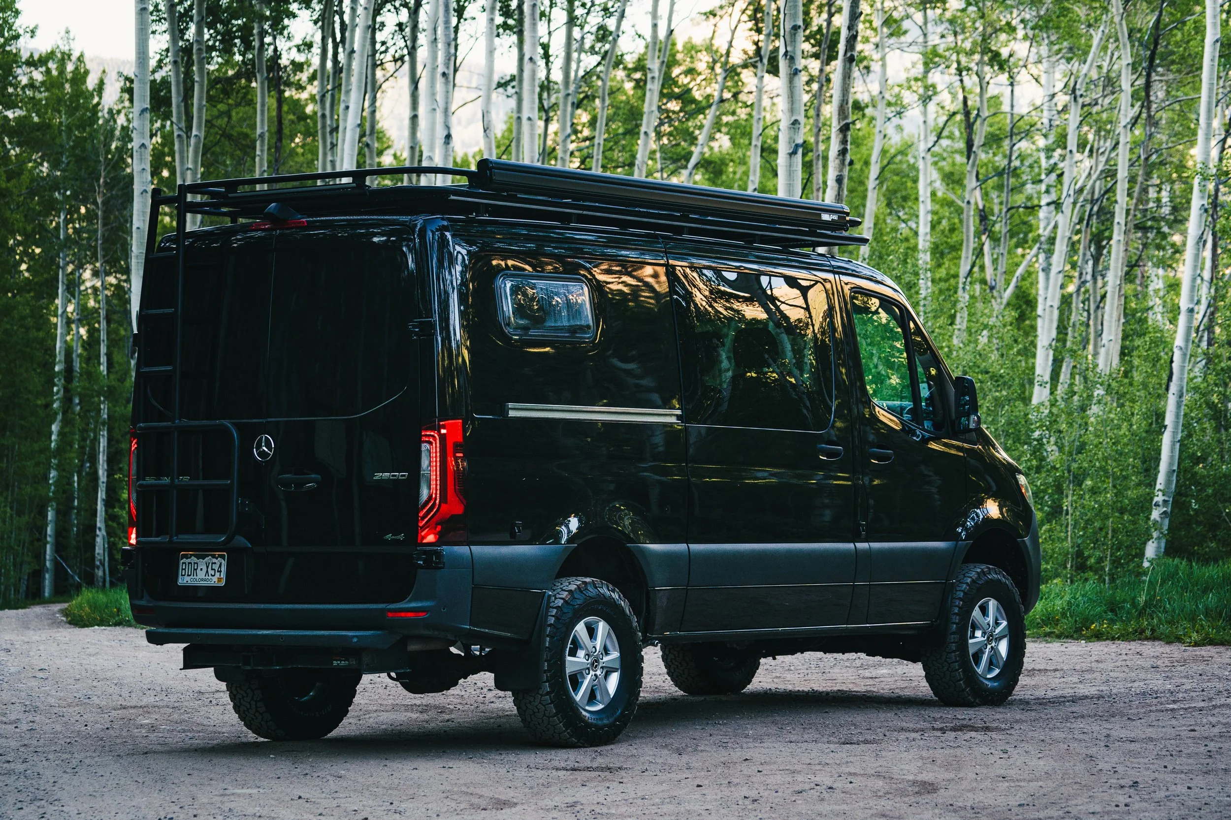 A black Mercedes-Benz camper van parked on a dirt path in a forest with tall, white-barked trees and green foliage.