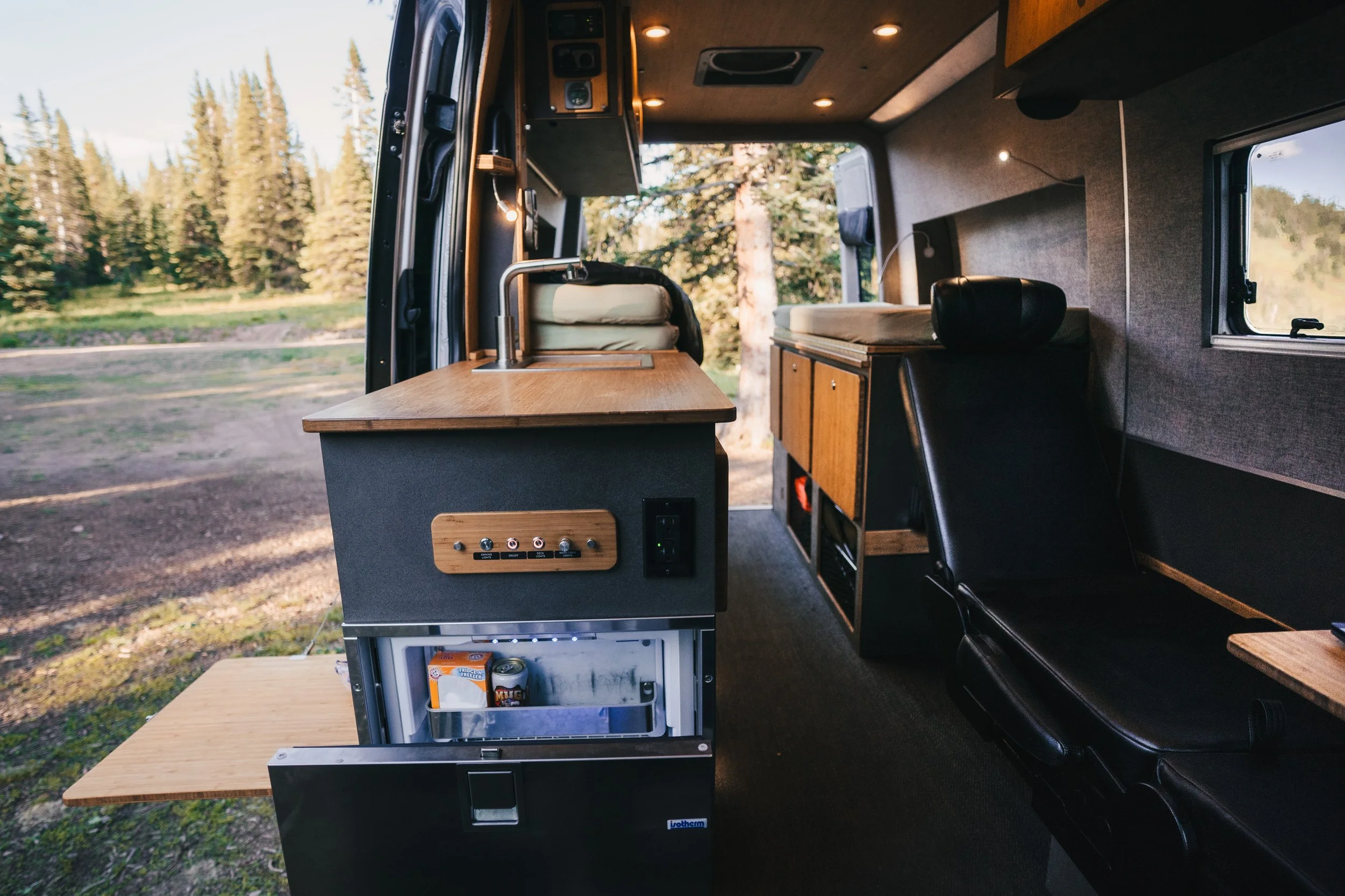 Interior of a converted camper van with a wooden counter, a small refrigerator, a black chair, and storage cabinets, with trees visible outside the window.